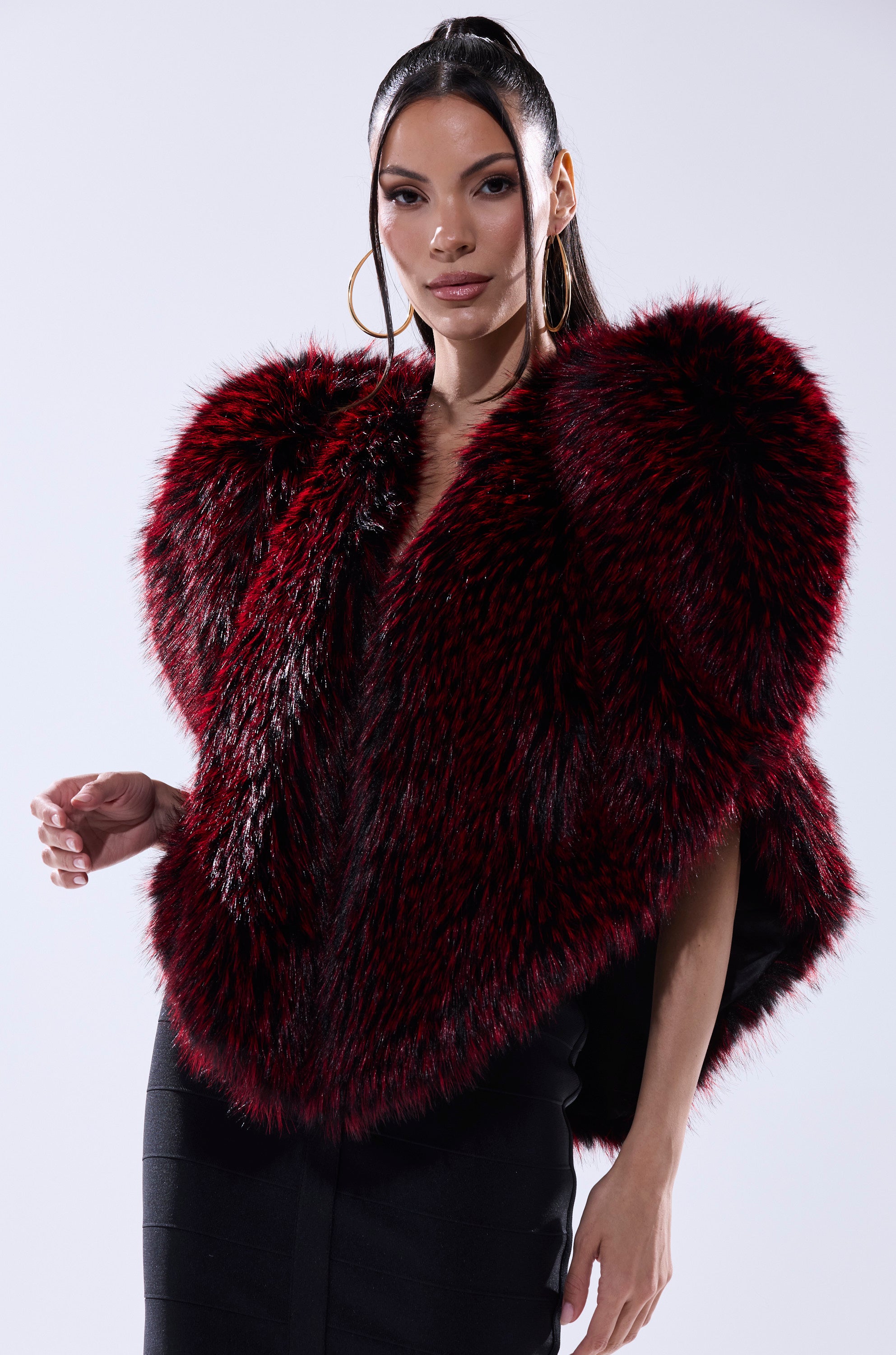 A woman confidently poses against a plain light background, wearing the STAR CROSSED LOVER FAUX FUR VEST in red over a fitted black dress.