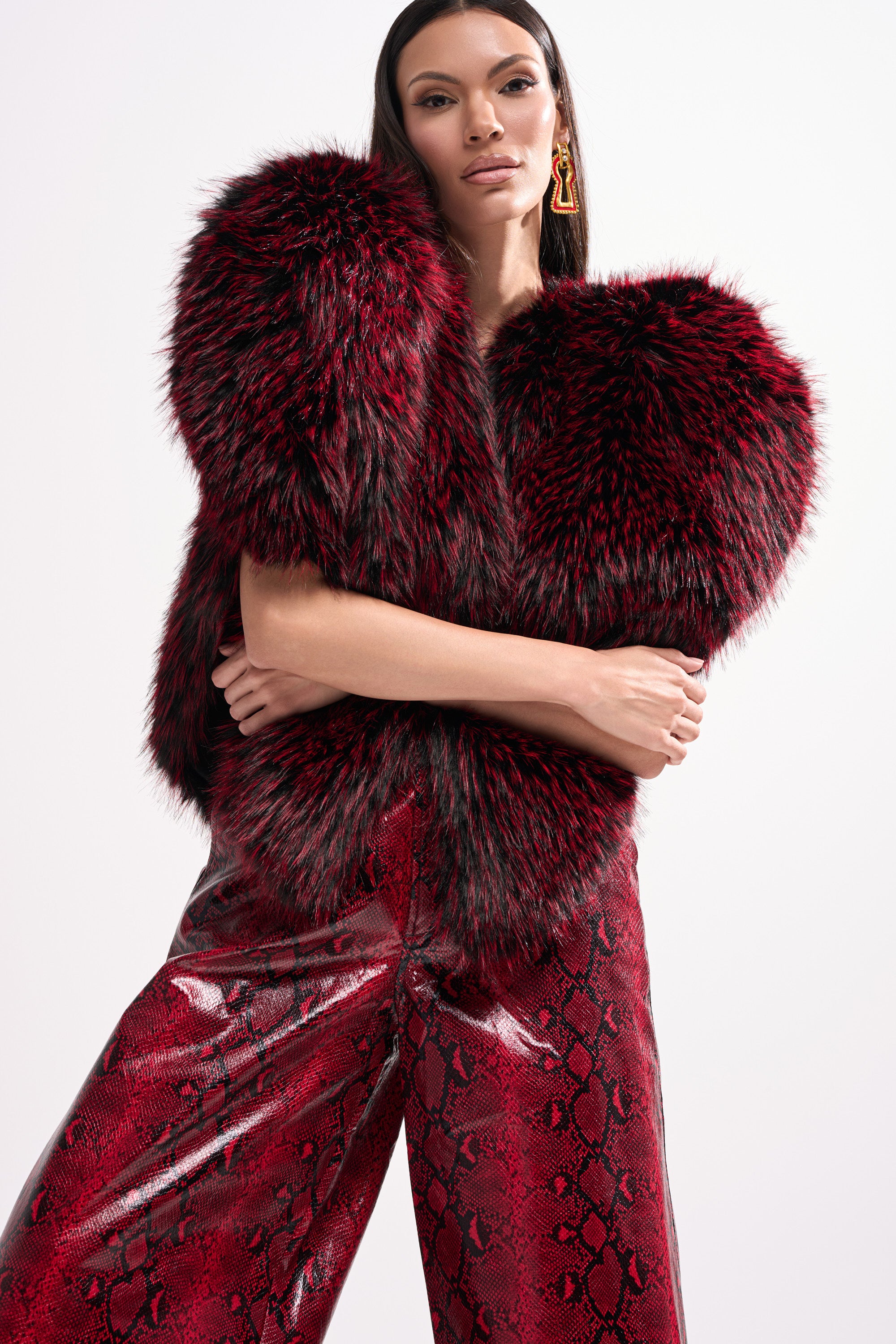 A woman poses confidently in the STAR CROSSED LOVER FAUX FUR VEST and shiny red snakeskin-patterned pants, accessorized with large statement earrings, against a plain light background.