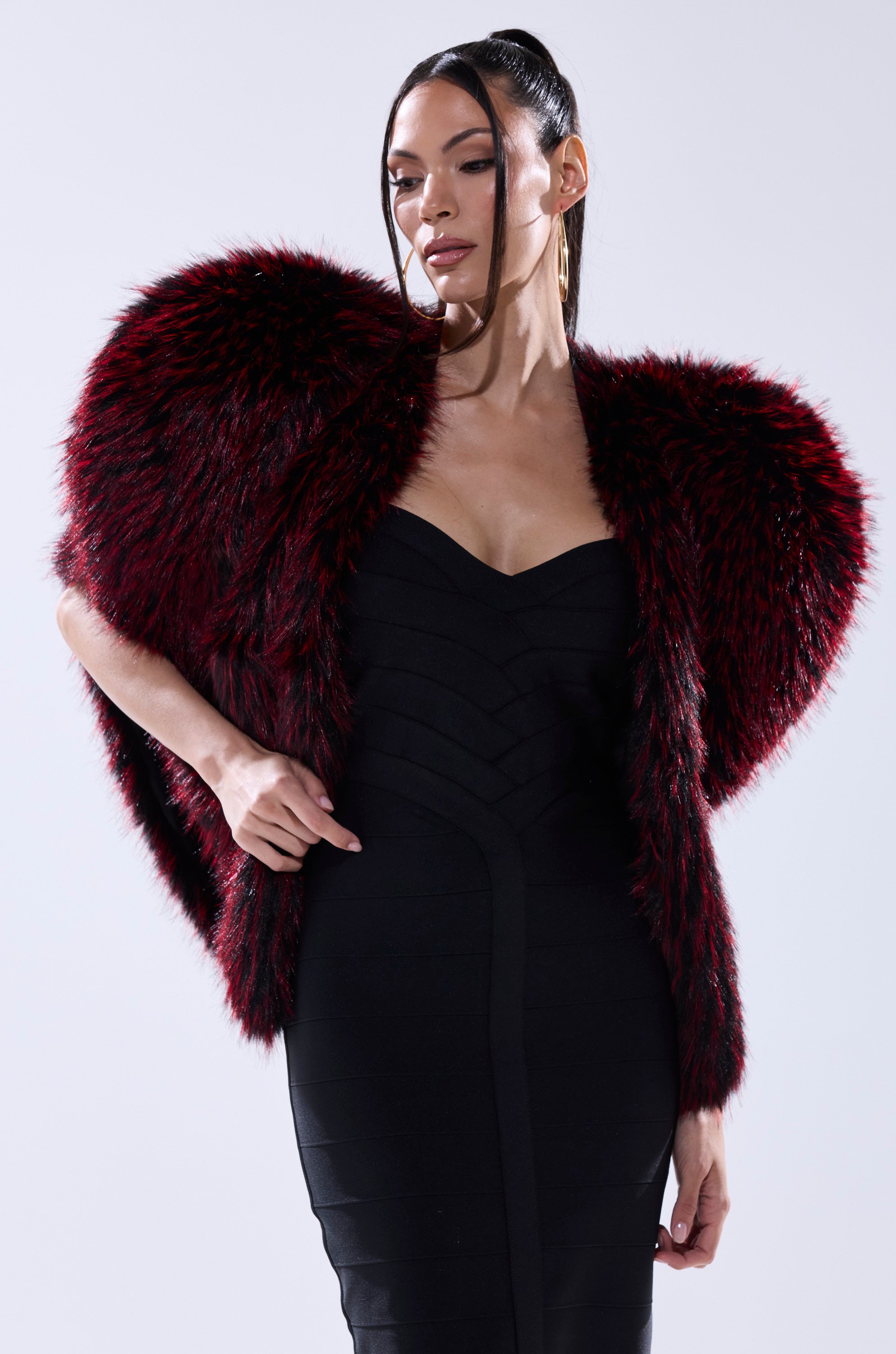 A woman poses confidently in a form-fitting black dress and the STAR CROSSED LOVER FAUX FUR VEST, a dramatic red-and-black piece from AKIRA Label, standing out boldly against a light gray background.