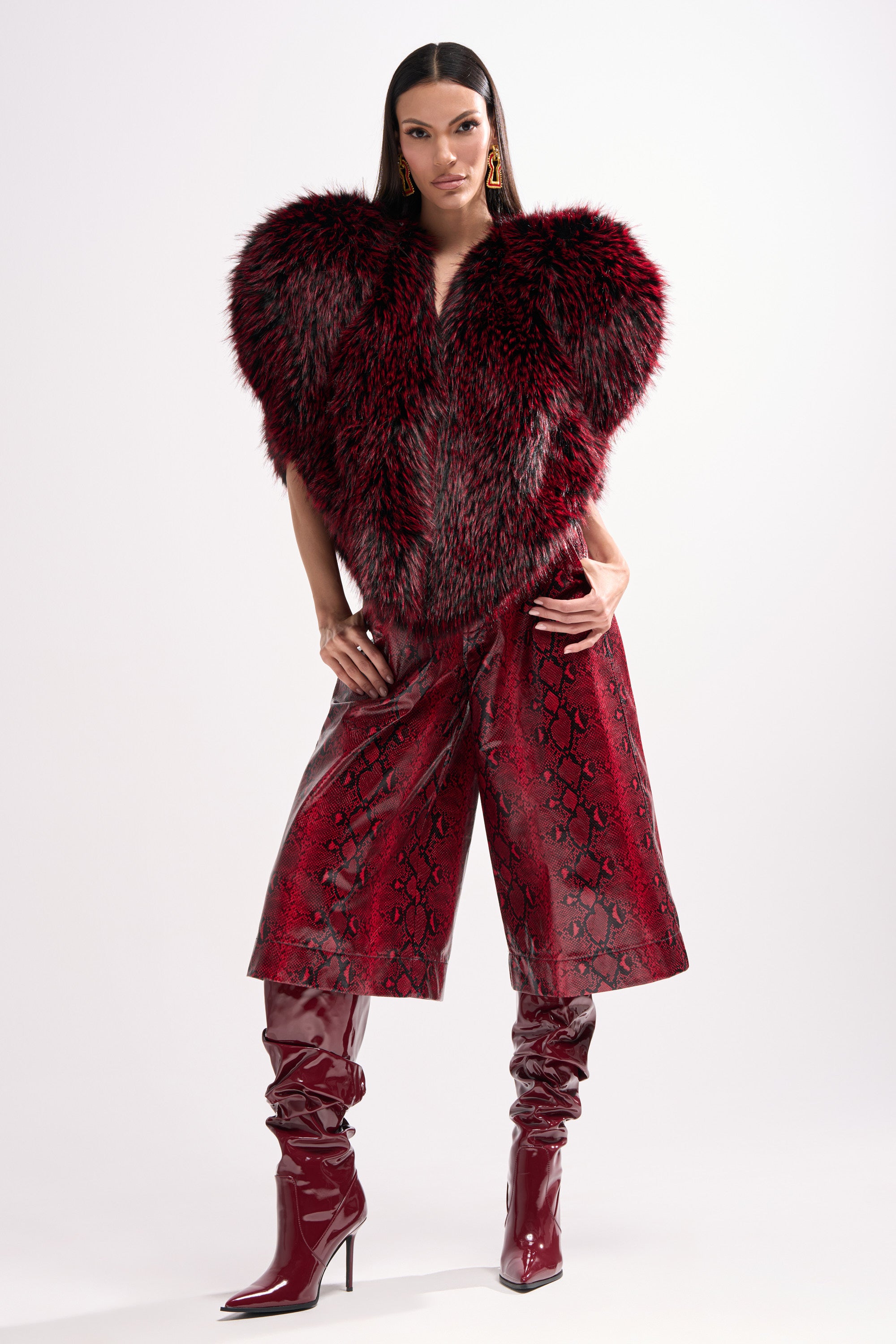 A woman stands confidently in a sleek STAR CROSSED LOVER FAUX FUR VEST, red snakeskin culotte pants, and shiny burgundy knee-high boots. Her straight hair and hands-on-hips pose contrast against a white background.
