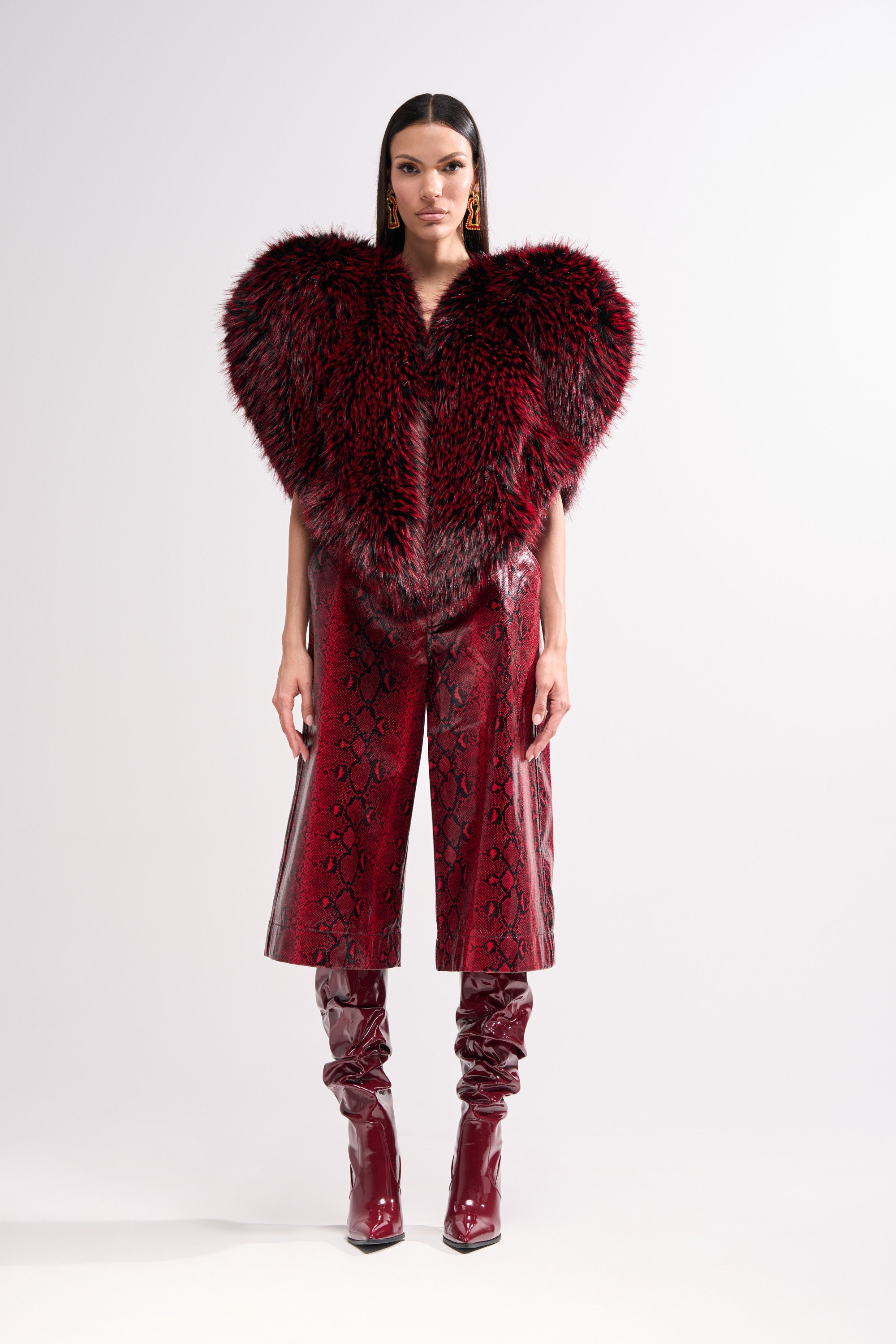 A woman stands against a plain background wearing the STAR CROSSED LOVER FAUX FUR VEST, red snakeskin-patterned wide-leg pants, shiny knee-high heeled boots, with straight hair and statement earrings.