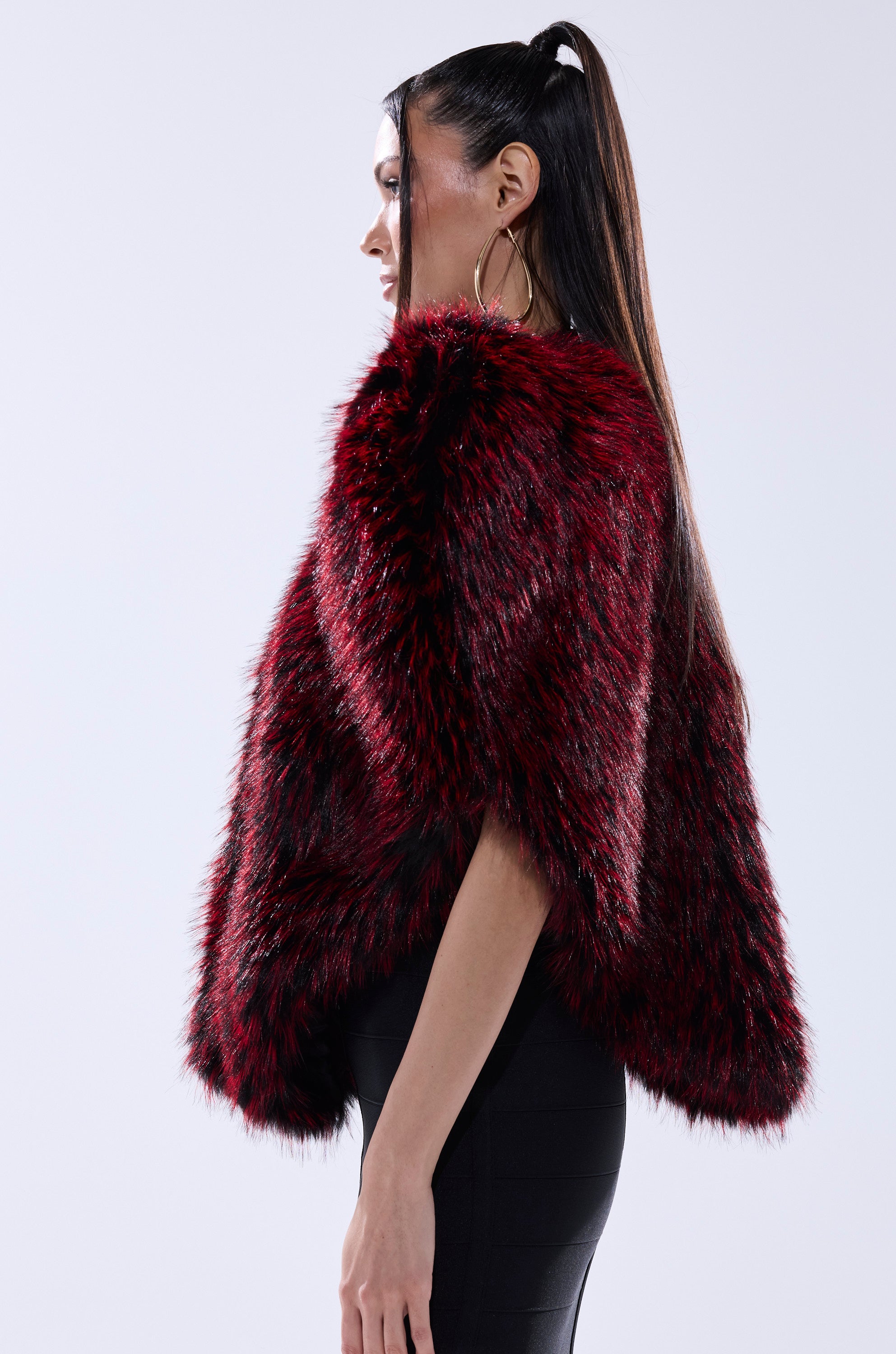 A woman with long, straight hair in a ponytail wears large hoop earrings and the STAR CROSSED LOVER FAUX FUR VEST—a bold, thick dark red and black layering piece—standing in profile against a plain light background.