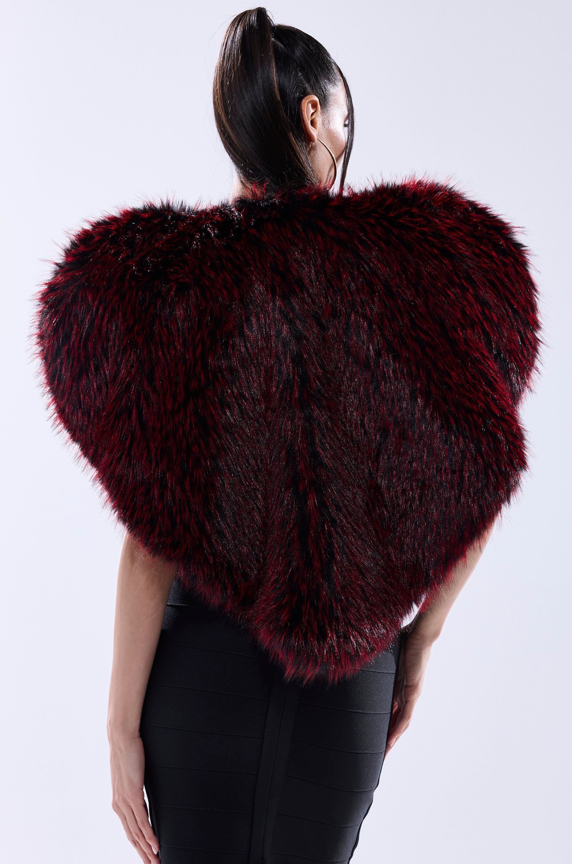 A woman wears the STAR CROSSED LOVER FAUX FUR VEST—a bold deep red and black heart-shaped piece—seen from behind with her hair in a high ponytail, paired with a fitted black skirt.
