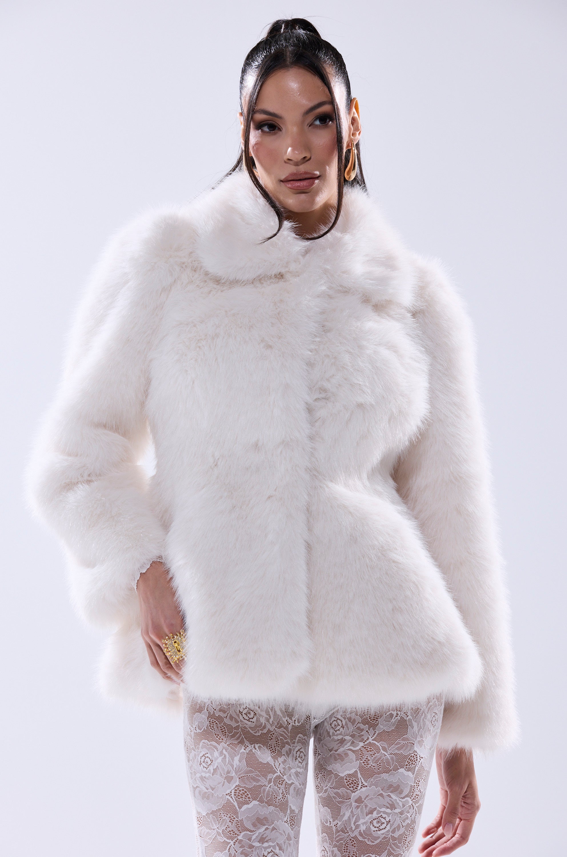 A person models the KENNY PEPLUM FAUX FUR jacket with a cinched waist and sheer white lace pants, standing confidently against a plain light background.