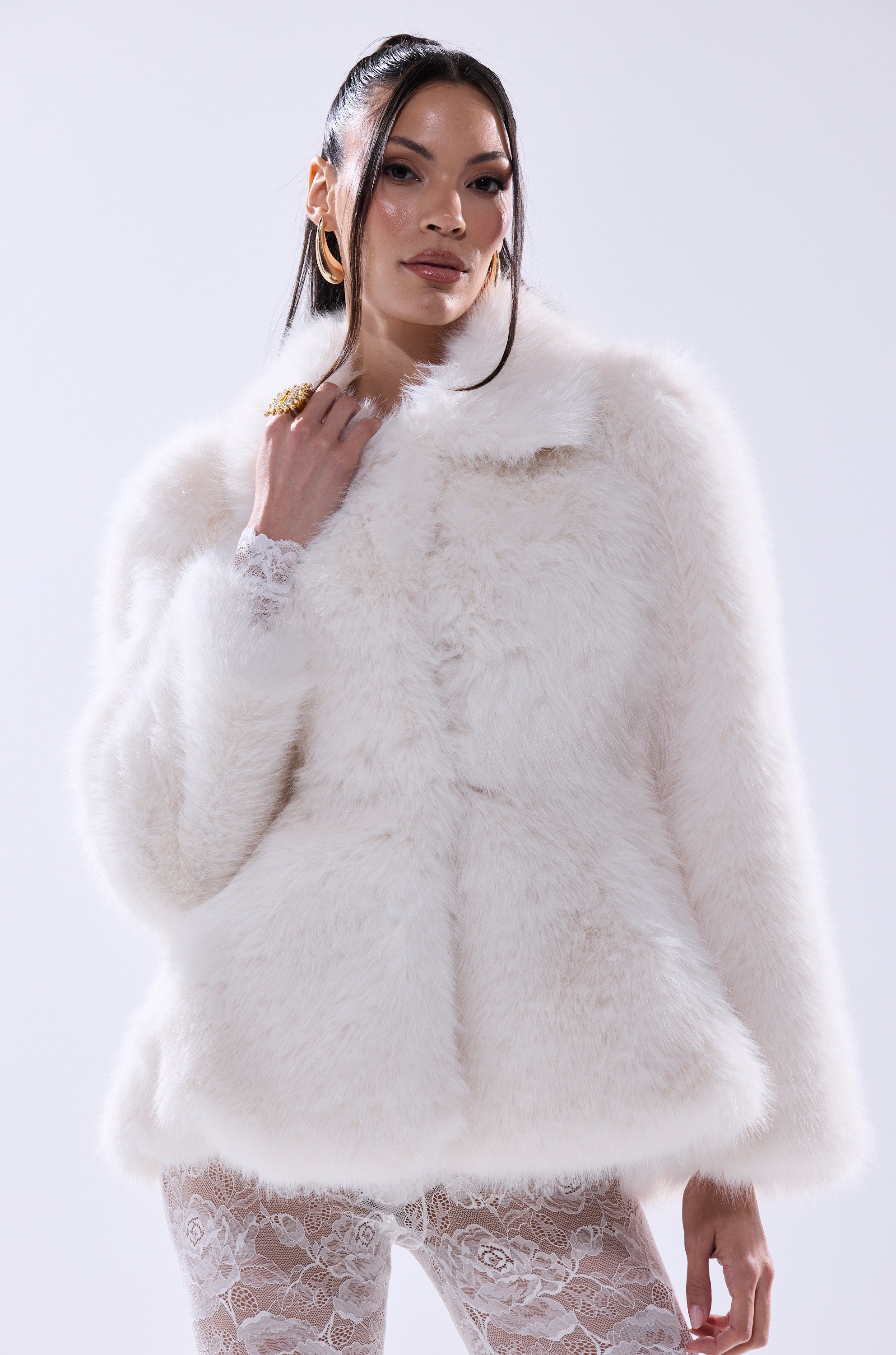 A woman models the KENNY PEPLUM FAUX FUR jacket featuring lace-trimmed sleeves and a cinched waist. She wears gold hoop earrings and rings, with long dark hair, standing against a plain light background.