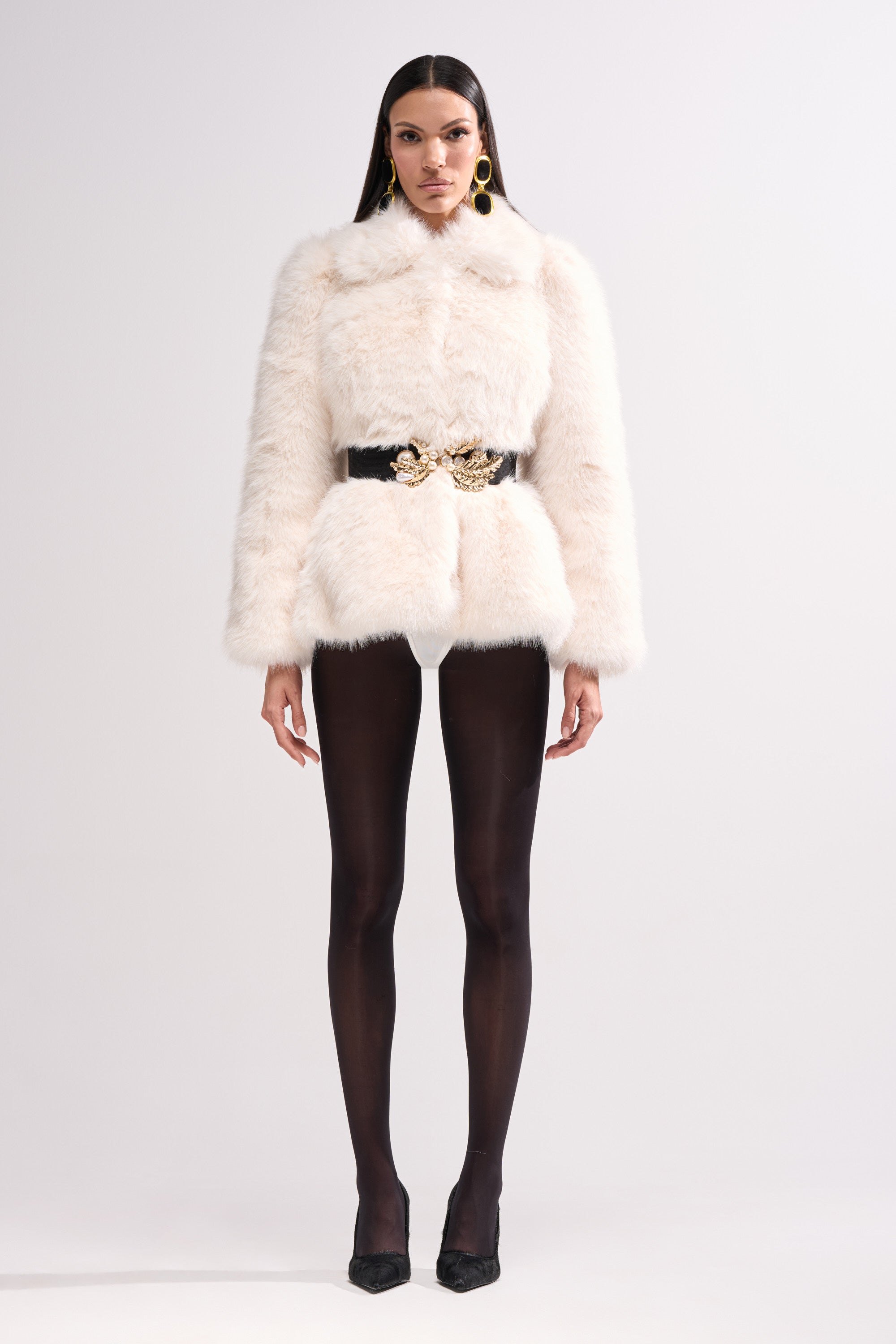 A woman poses against a plain background wearing the KENNY PEPLUM FAUX FUR jacket, paired with black sheer tights, black heels, and large gold earrings.