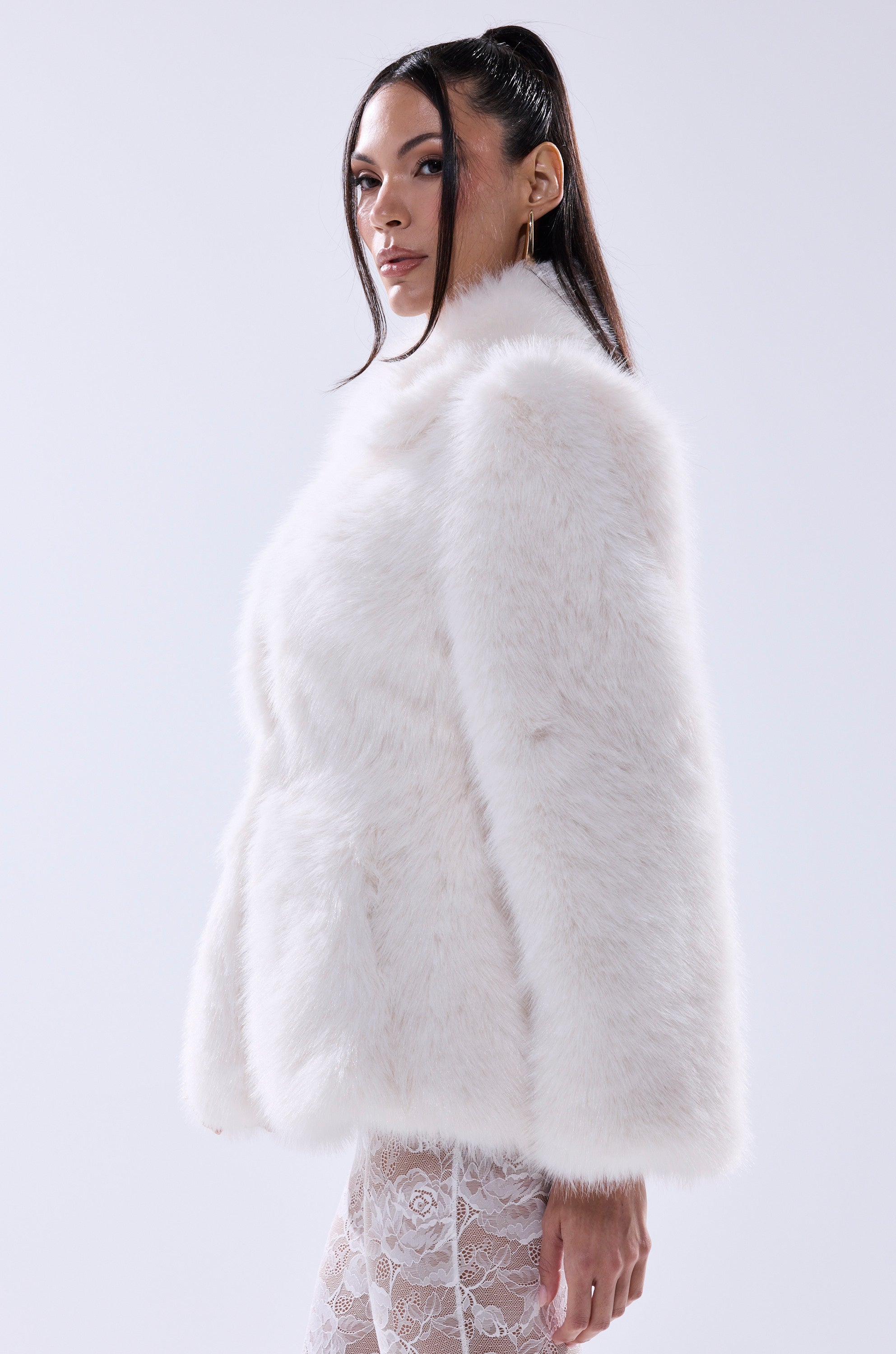 A woman with long dark hair wears the KENNY PEPLUM FAUX FUR jacket and sheer lace pants, standing sideways and looking at the camera against a plain light background.