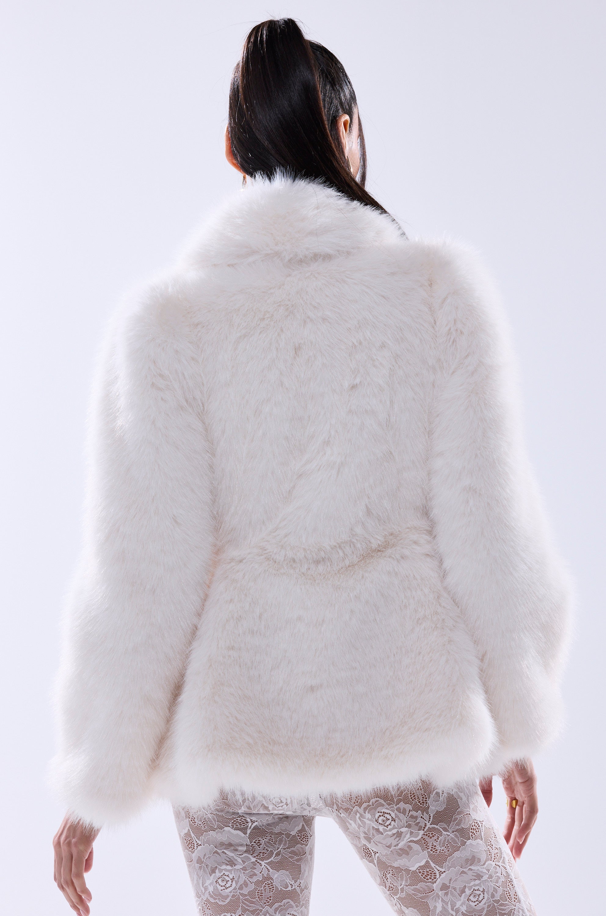 A woman with long dark hair in a high ponytail is seen from behind wearing the KENNY PEPLUM FAUX FUR jacket and sheer lace-patterned leggings, highlighting trendy women's outerwear against a simple light background.