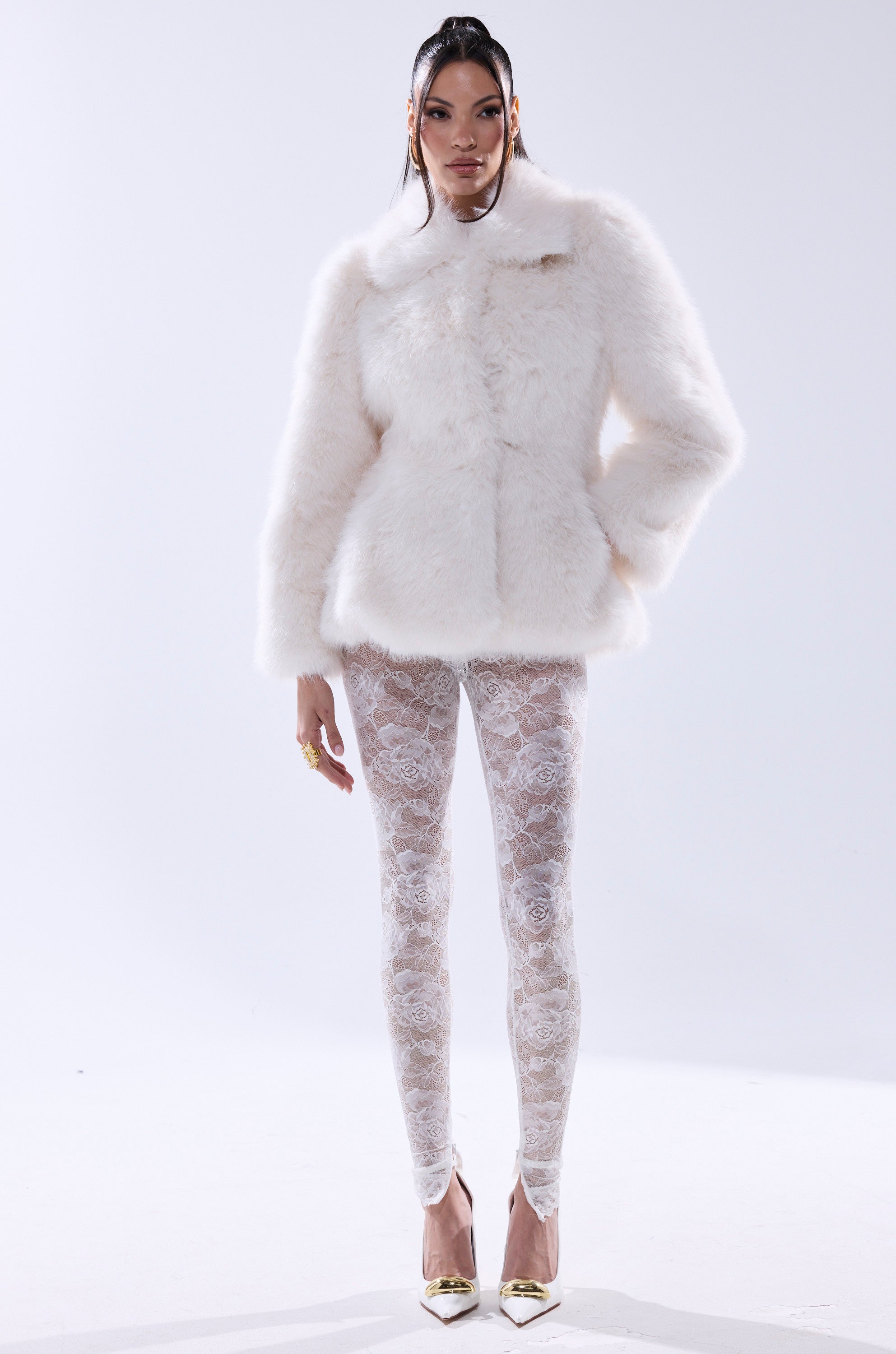 A woman poses against a plain light background in the KENNY PEPLUM FAUX FUR jacket, styled with white lace leggings, gold pointed-toe heels, drop earrings, and sleek straight hair.
