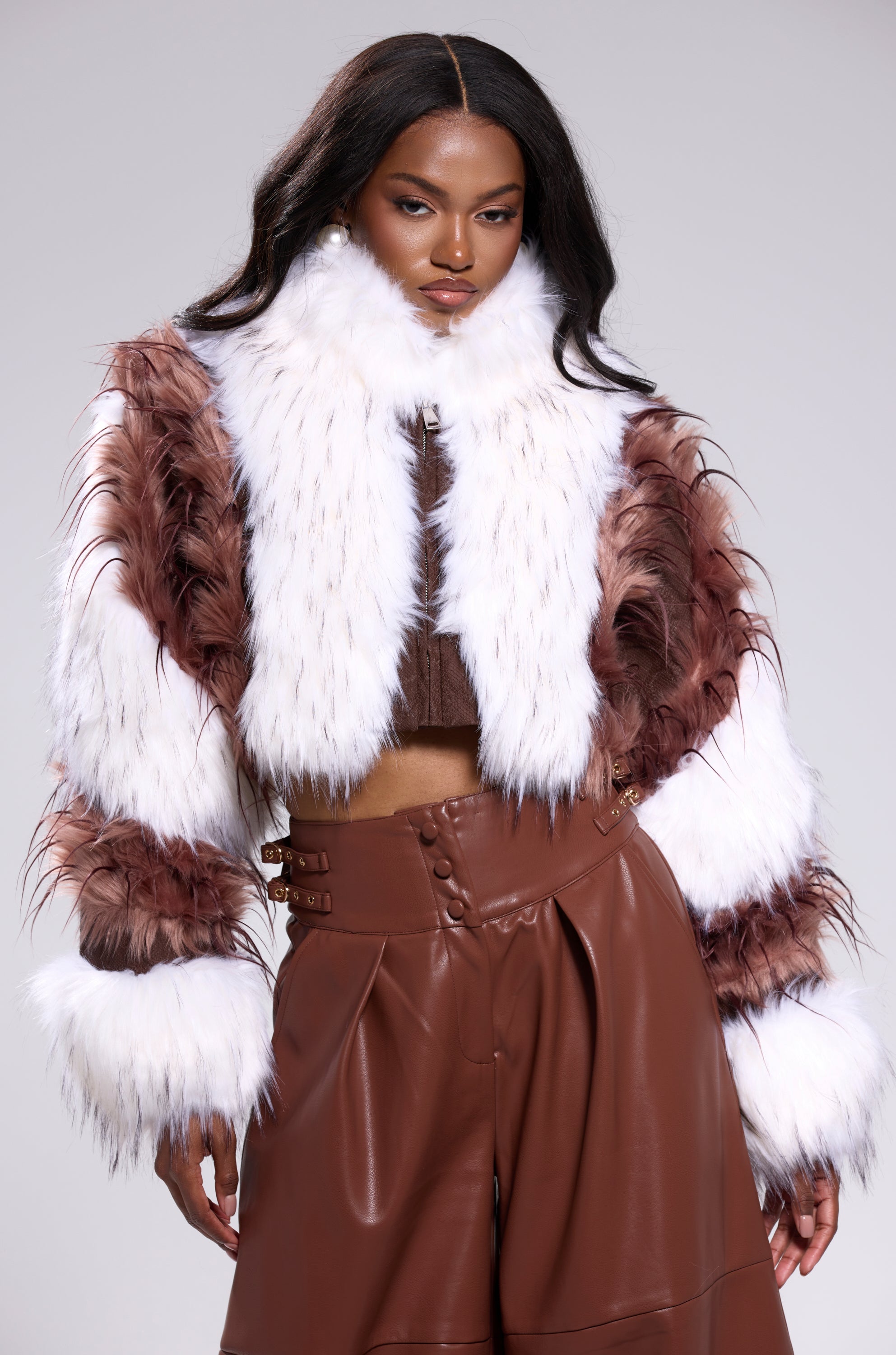 A woman with long, wavy black hair poses confidently in the FRANKIE PATCHWORK FAUX FUR jacket, featuring a high collar and paired with high-waisted brown leather pants against a plain light gray background. 0