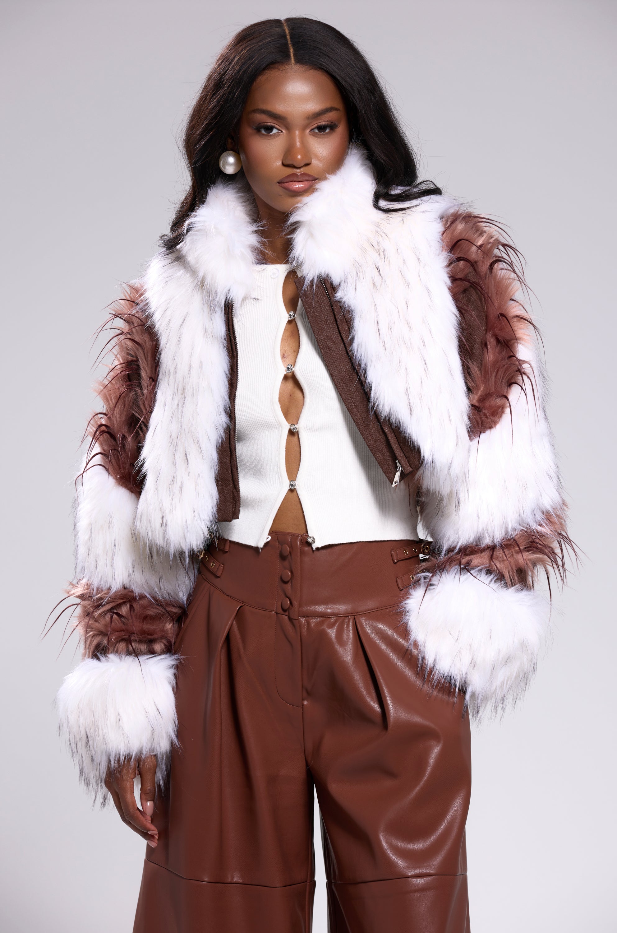 A woman poses confidently in a white top, high-waisted brown leather wide-leg pants, and the FRANKIE PATCHWORK FAUX FUR jacket against a light gray background. 1