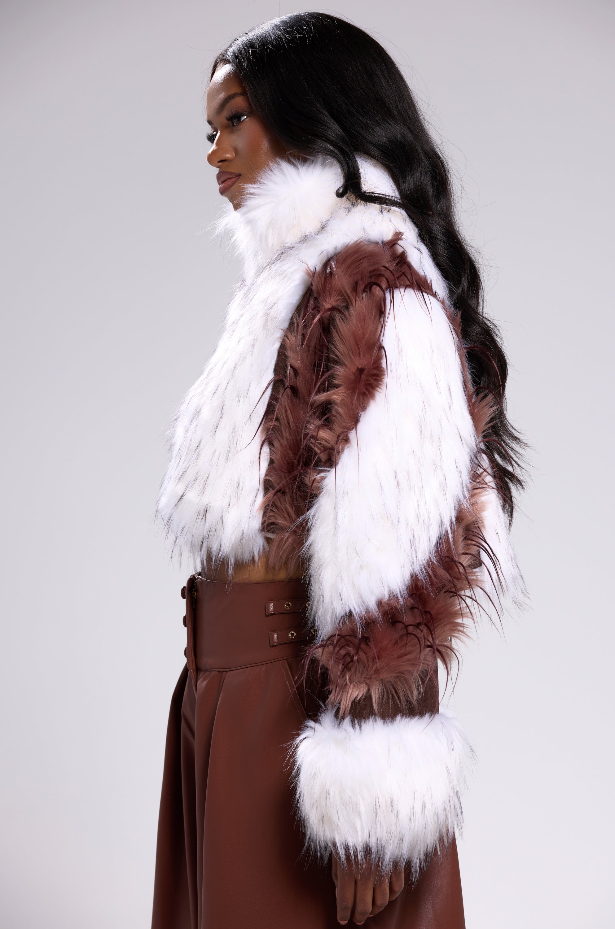 A woman with long, wavy black hair models the FRANKIE PATCHWORK FAUX FUR jacket, featuring feathered details, paired with high-waisted brown pants against a plain light background. 2