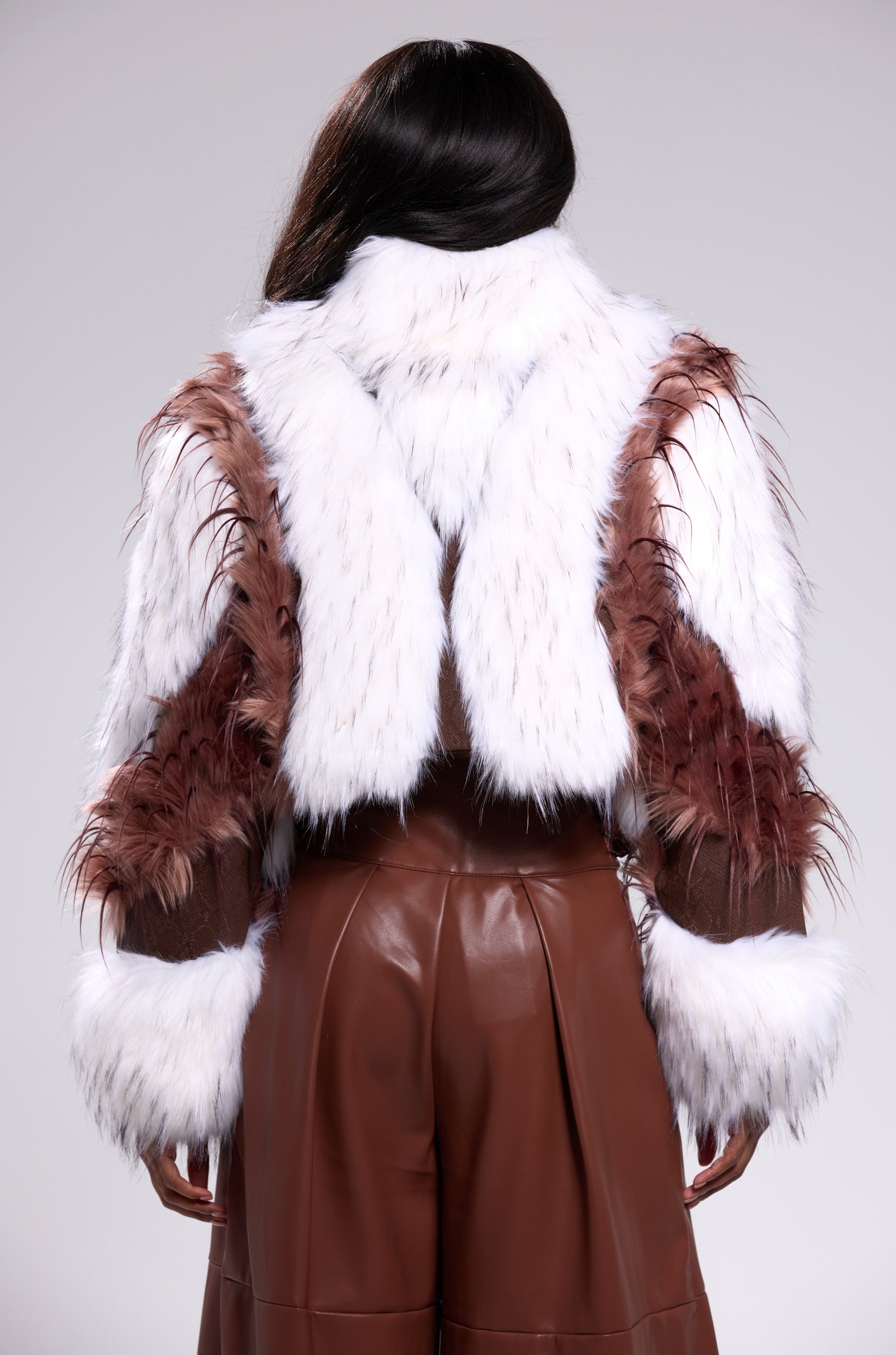 A person faces away from the camera, wearing the FRANKIE PATCHWORK FAUX FUR jacket and high-waisted brown leather pants, set against a neutral background. 3