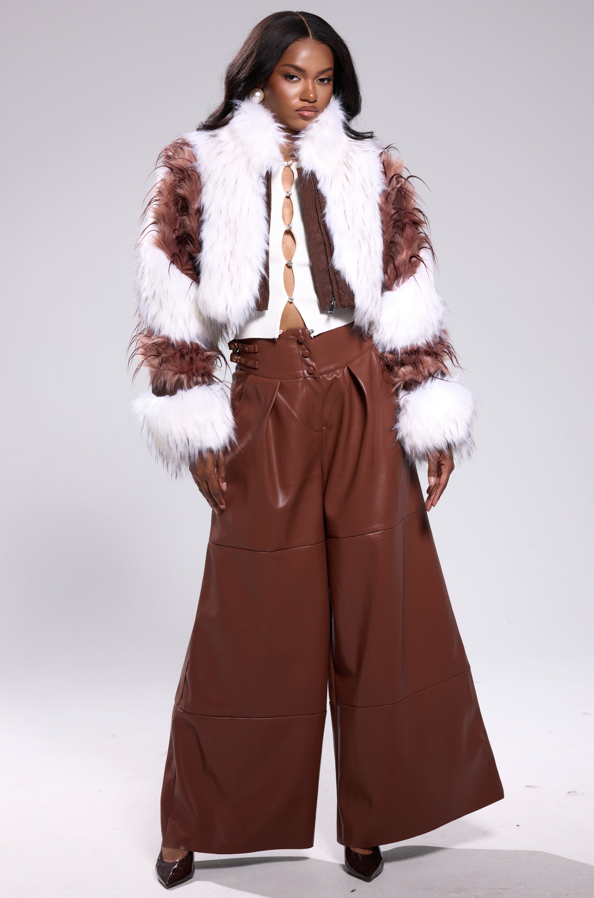 A woman poses confidently in the FRANKIE PATCHWORK FAUX FUR jacket, wide-legged brown leather pants, a white cut-out top, and brown heels against a plain gray background. 5