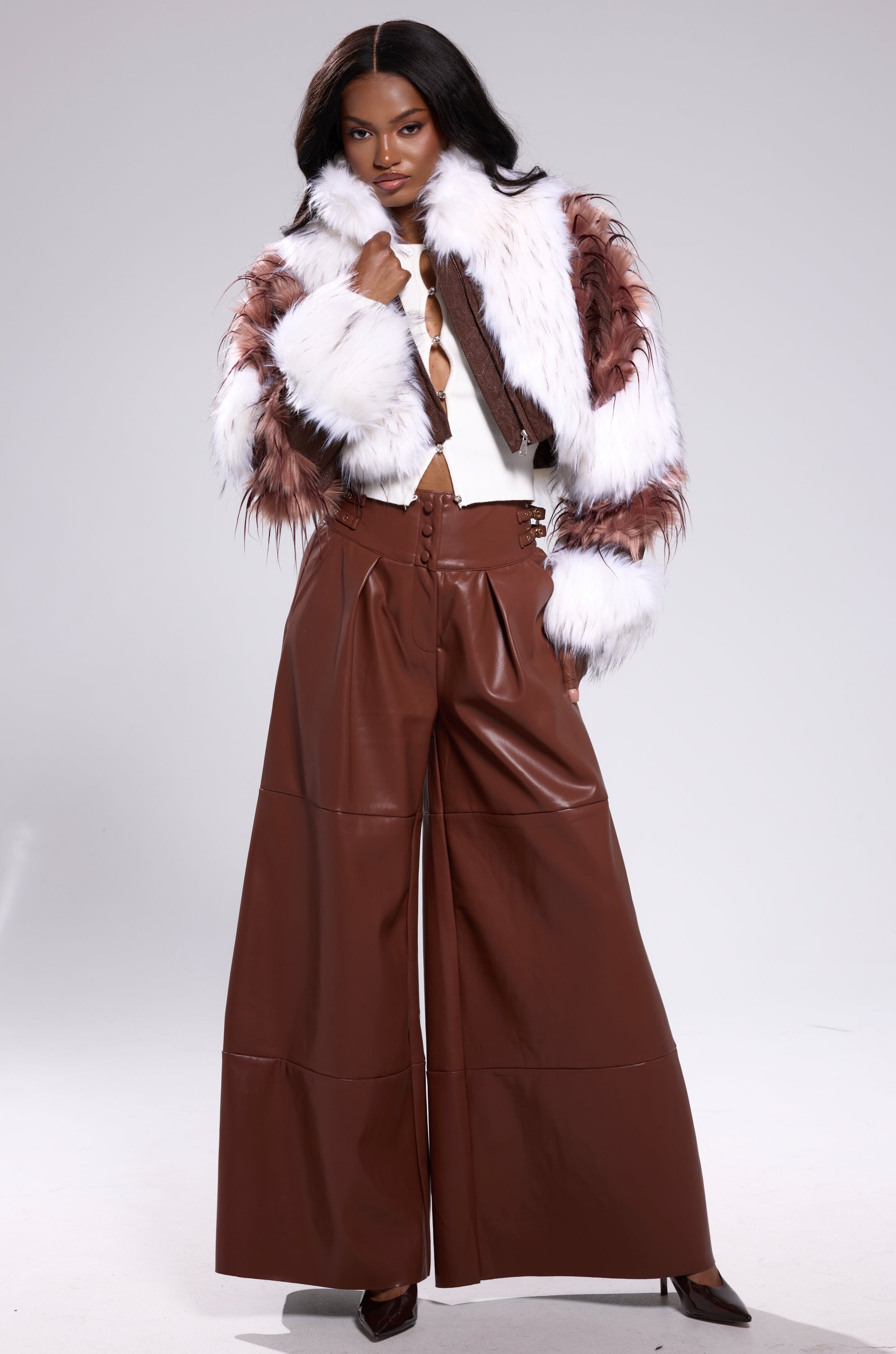 A woman poses confidently in wide-leg brown leather pants, a white top, and the FRANKIE PATCHWORK FAUX FUR jacket with white and brown accents against a plain light background. 6