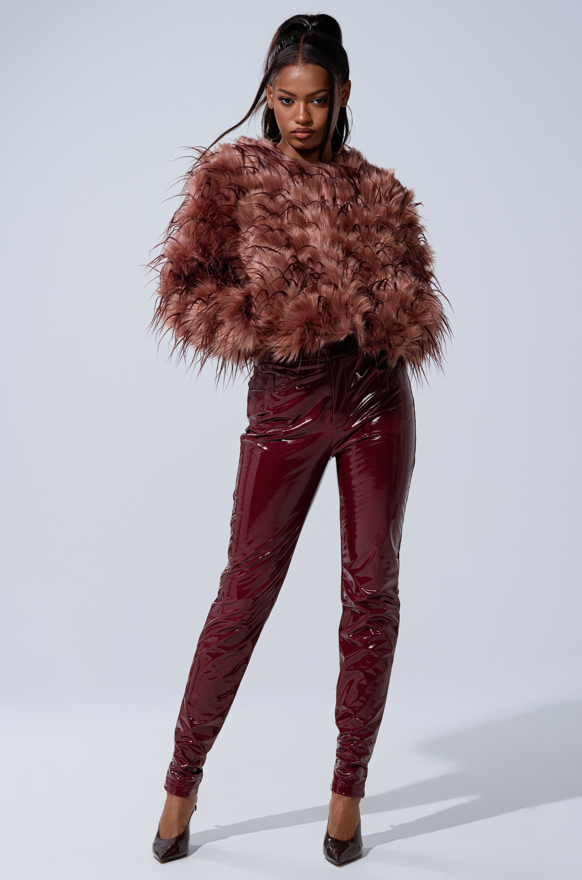 A woman stands confidently in the PIPER TEXTURED FAUX FUR COAT and shiny burgundy vinyl pants with matching pointed heels against a plain light background.