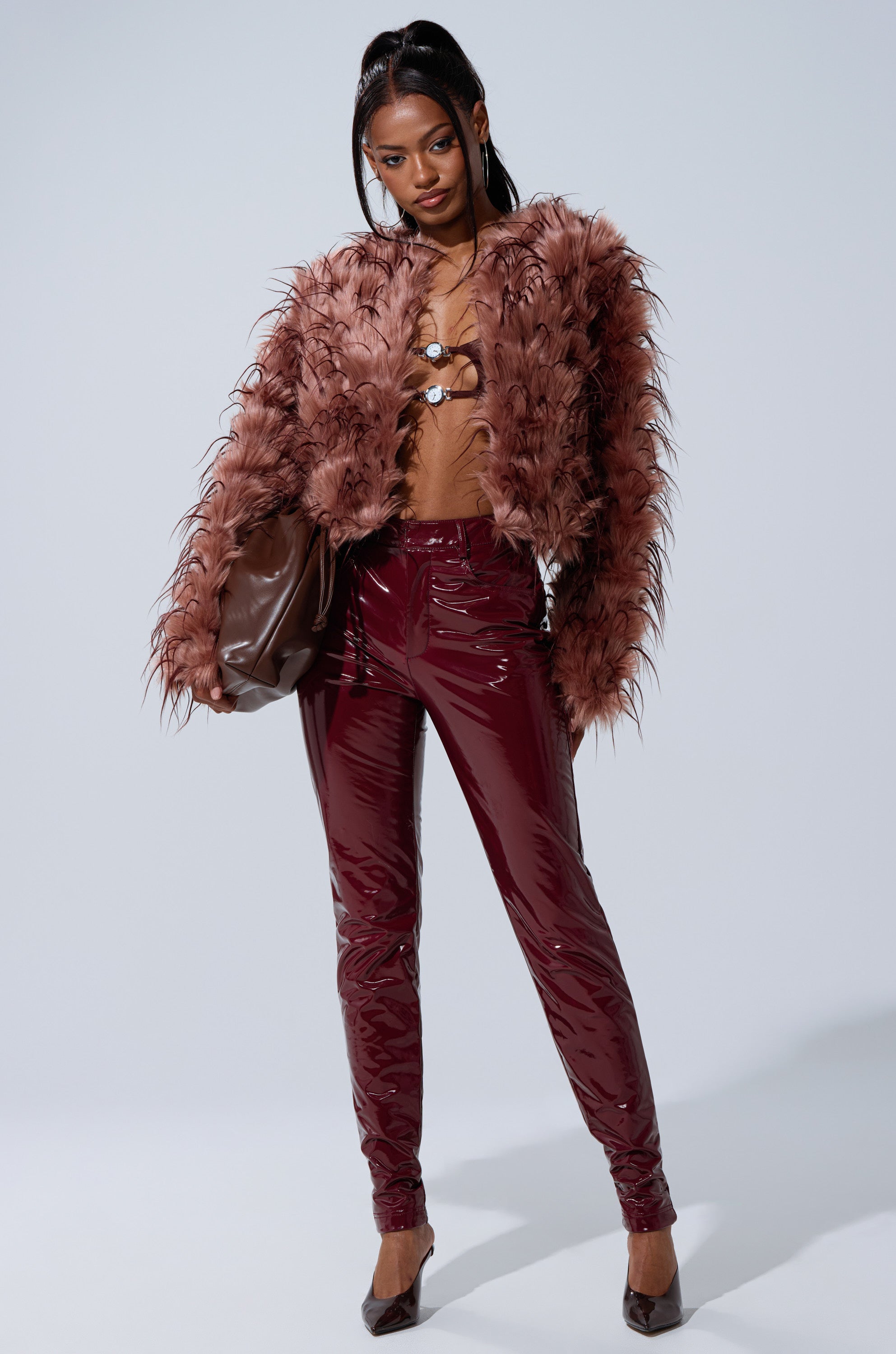 A woman poses confidently in the PIPER TEXTURED FAUX FUR COAT, styled with shiny burgundy pants, pointed heels, a large brown clutch, and her hair slicked back with two face-framing strands.
