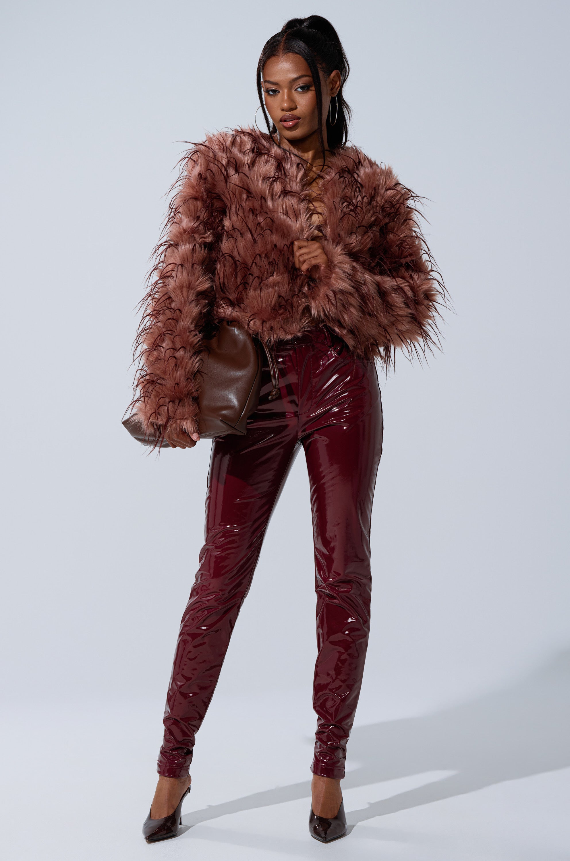 A woman poses confidently in the PIPER TEXTURED FAUX FUR COAT with shiny burgundy vinyl pants, pointed heels, a brown shoulder bag, and a sleek ponytail. The background is plain white.
