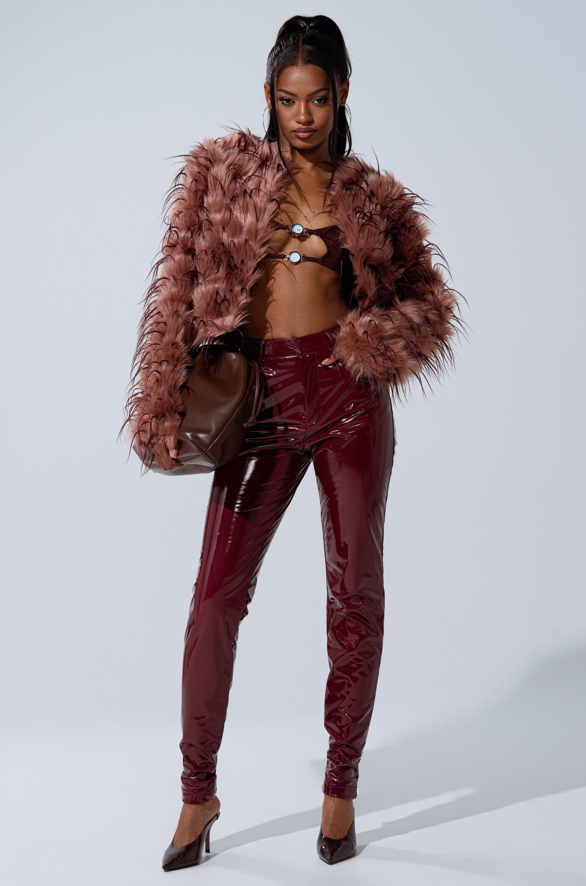 A woman poses confidently in the PIPER TEXTURED FAUX FUR COAT with maroon vinyl pants, brown heels, and a large brown handbag. Her hair is styled in two high pigtails against a plain, light-colored background.