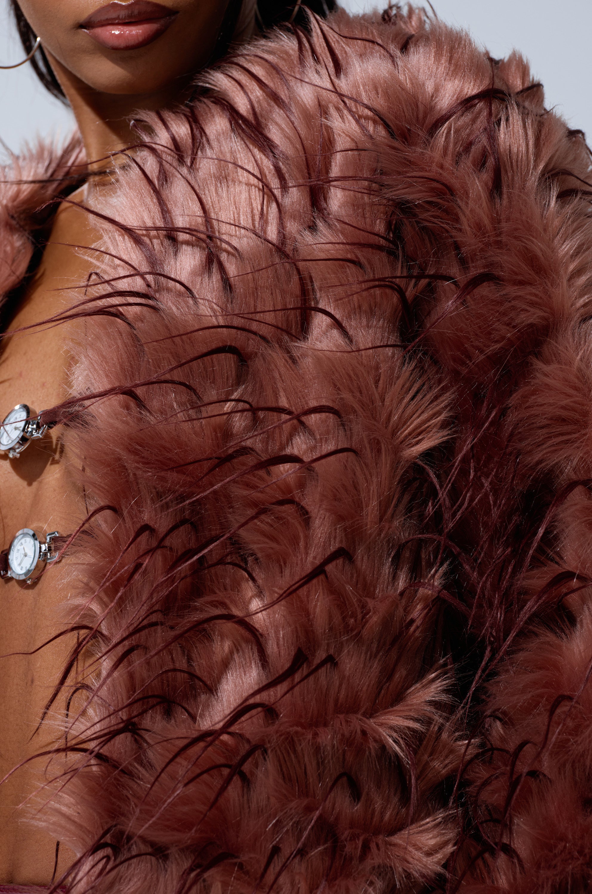 A close-up of a person wearing the PIPER TEXTURED FAUX FUR COAT in mauve, featuring long, dark accents. Only part of their face and chest is visible, accessorized with two small watch pendants.