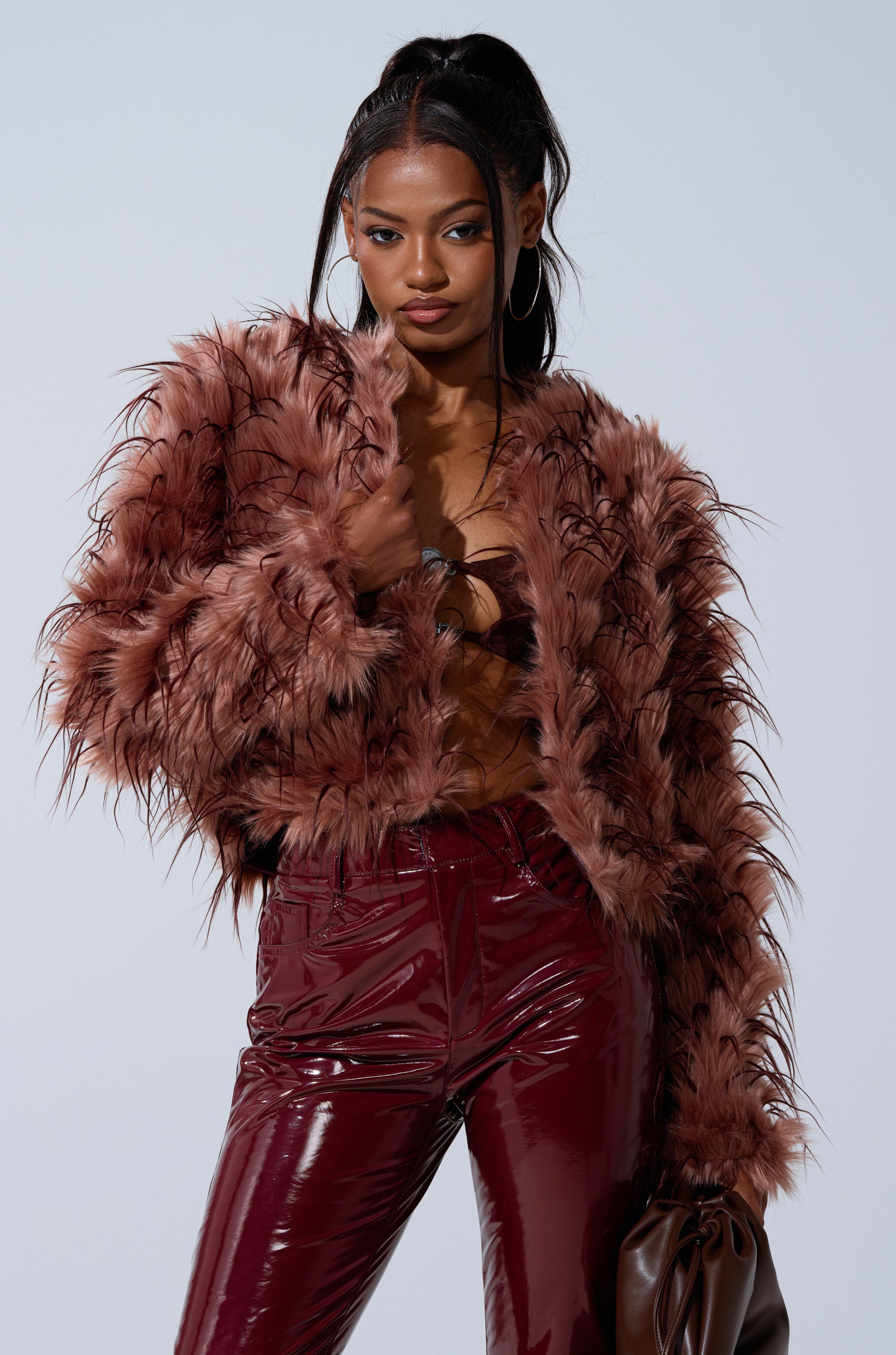 A woman poses confidently in the PIPER TEXTURED FAUX FUR COAT, styled with shiny burgundy pants and a brown bag. She wears her long hair in a high ponytail and large hoop earrings against a plain, light background.