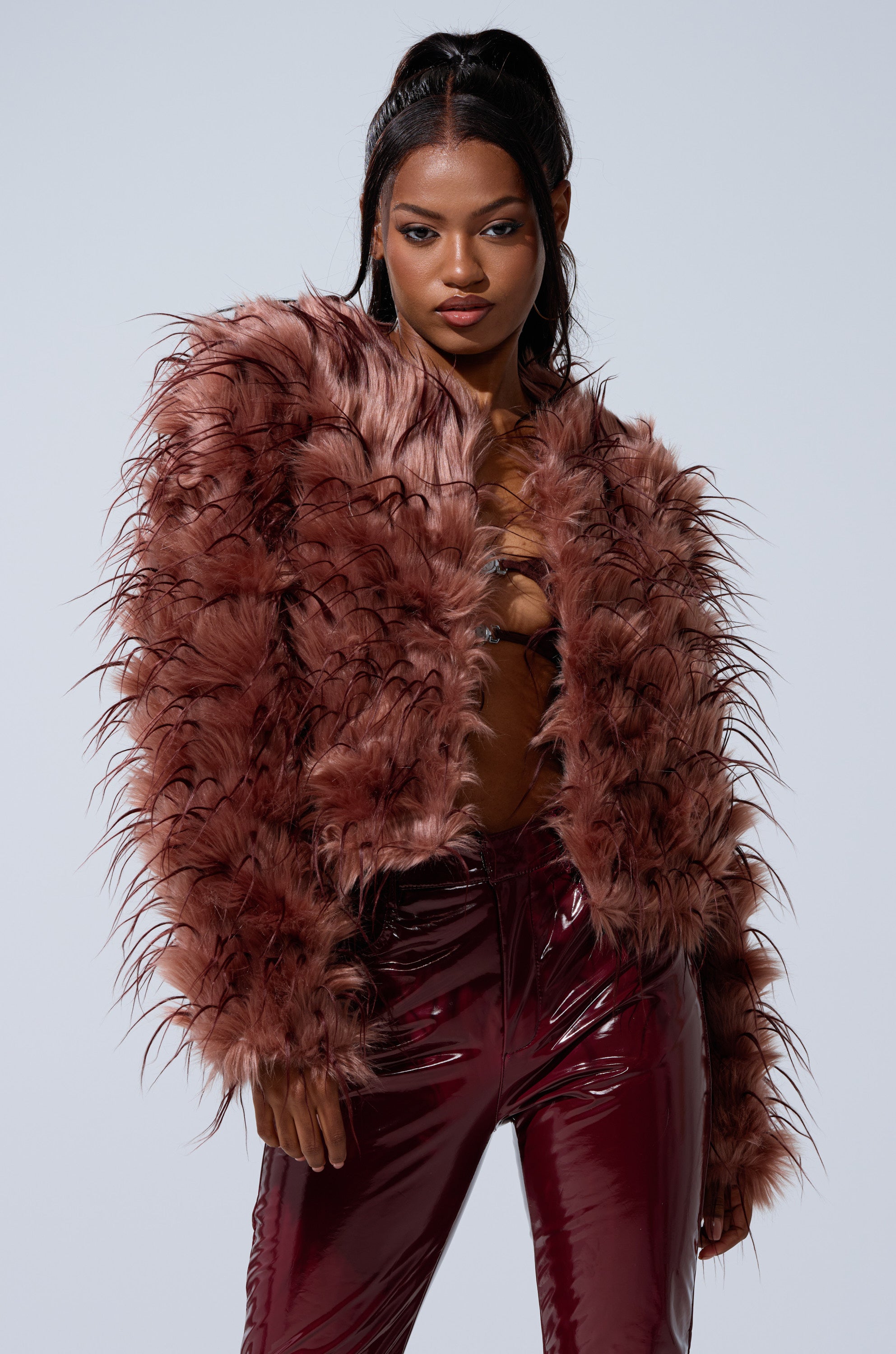 A woman poses confidently against a plain light gray background, wearing the PIPER TEXTURED FAUX FUR COAT in maroon with a boxy fit and glossy maroon pants.