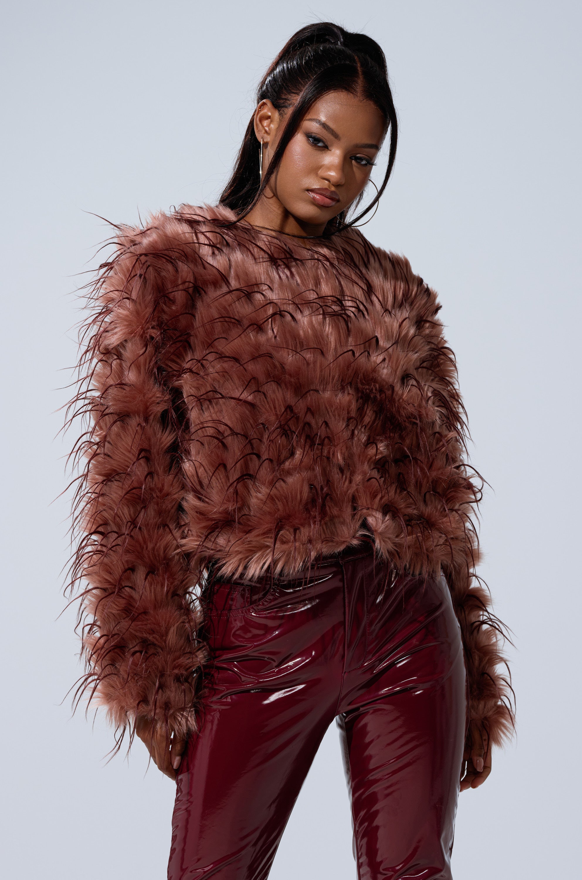 A woman models the PIPER TEXTURED FAUX FUR COAT in dark mauve, styled with shiny maroon pants, posing against a plain light background and gazing slightly to the side with a neutral expression.