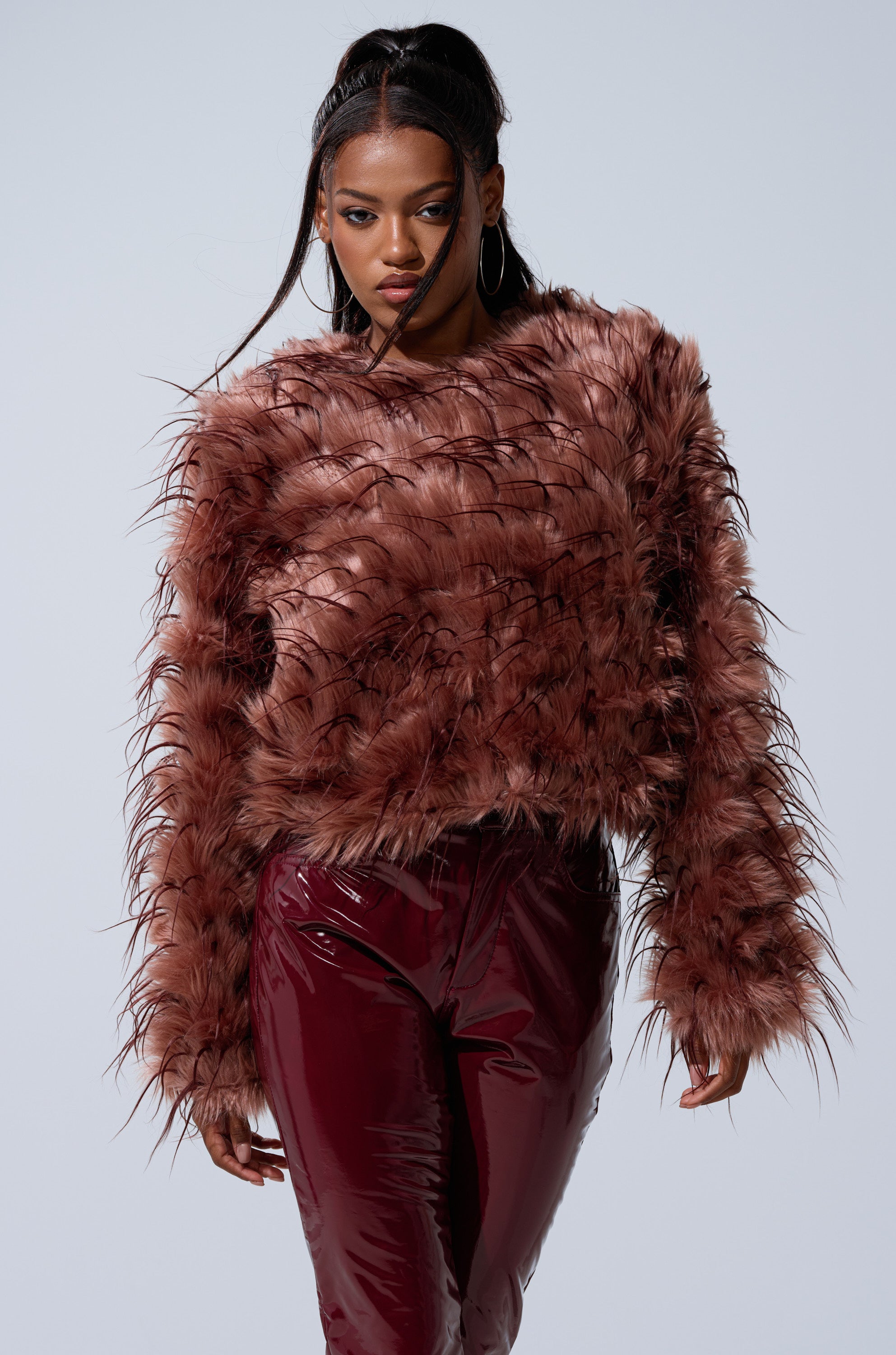 A woman wearing the PIPER TEXTURED FAUX FUR COAT and shiny burgundy pants stands against a plain light background, looking forward with a confident expression.