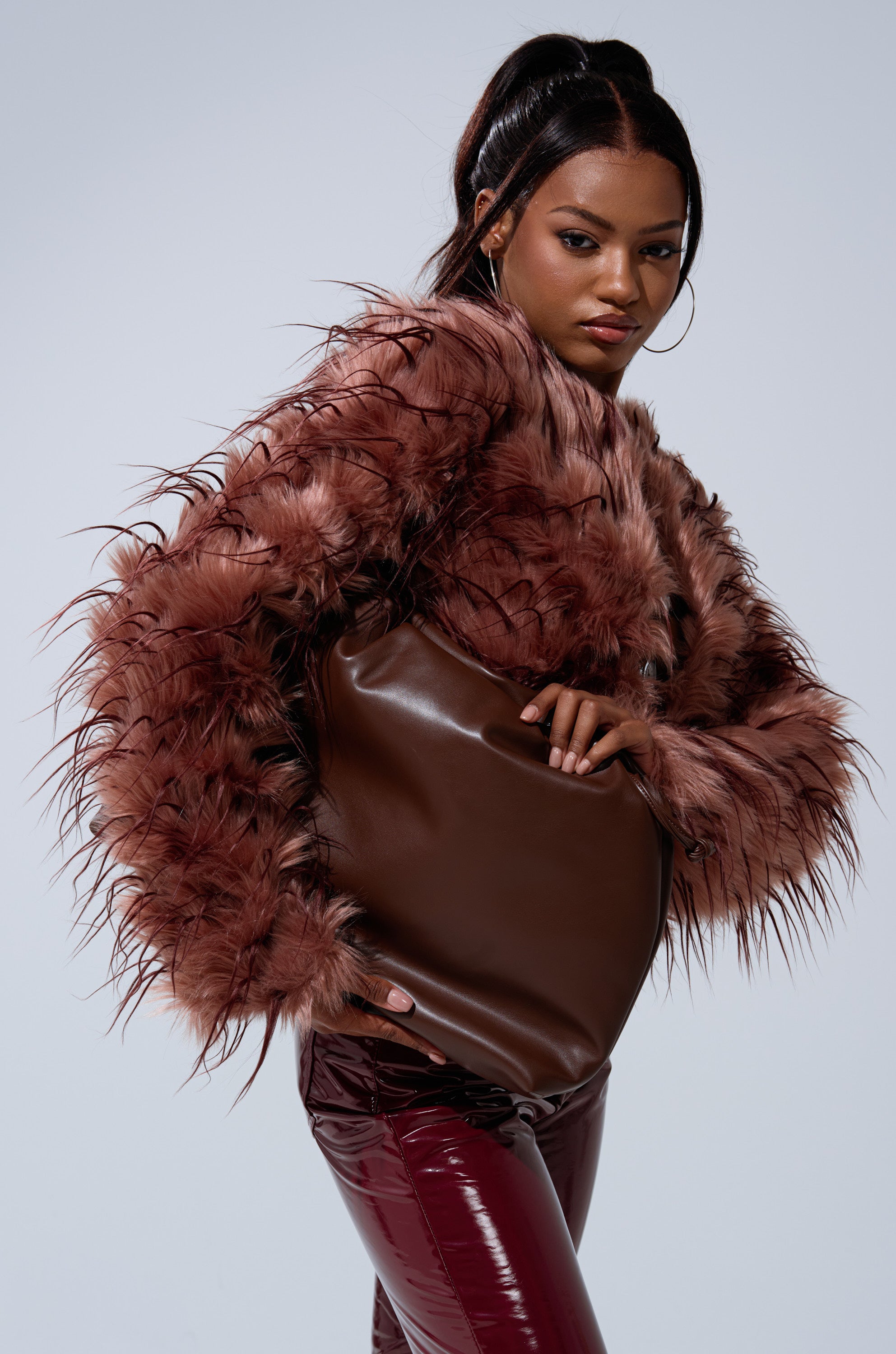 A woman in a PIPER TEXTURED FAUX FUR COAT and shiny burgundy pants poses confidently with a large brown handbag against a plain light background.