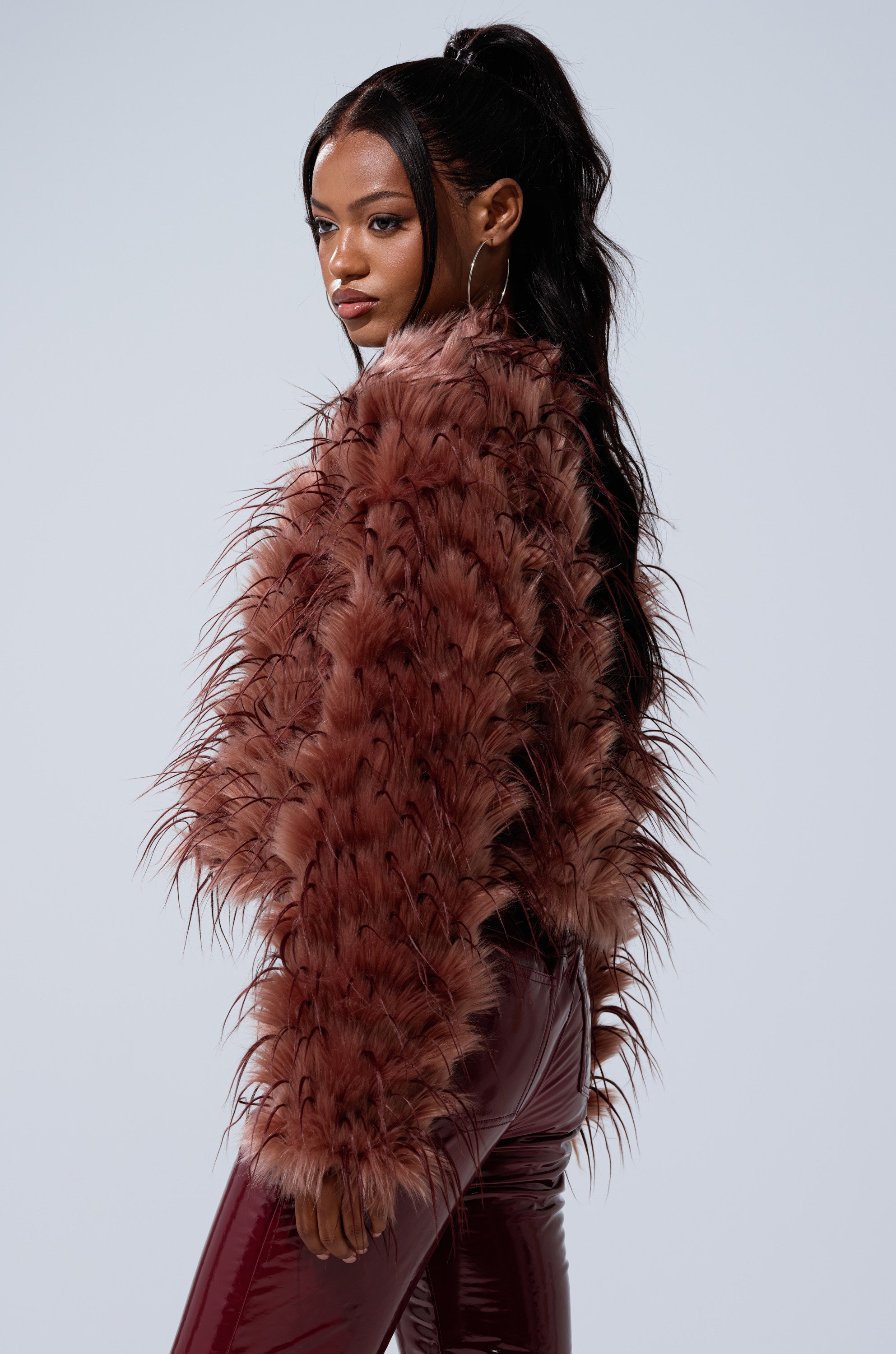 A woman models the PIPER TEXTURED FAUX FUR COAT with shiny maroon pants. Her long, wavy dark hair and large hoop earrings complete the look as she gazes sideways against a plain light background.