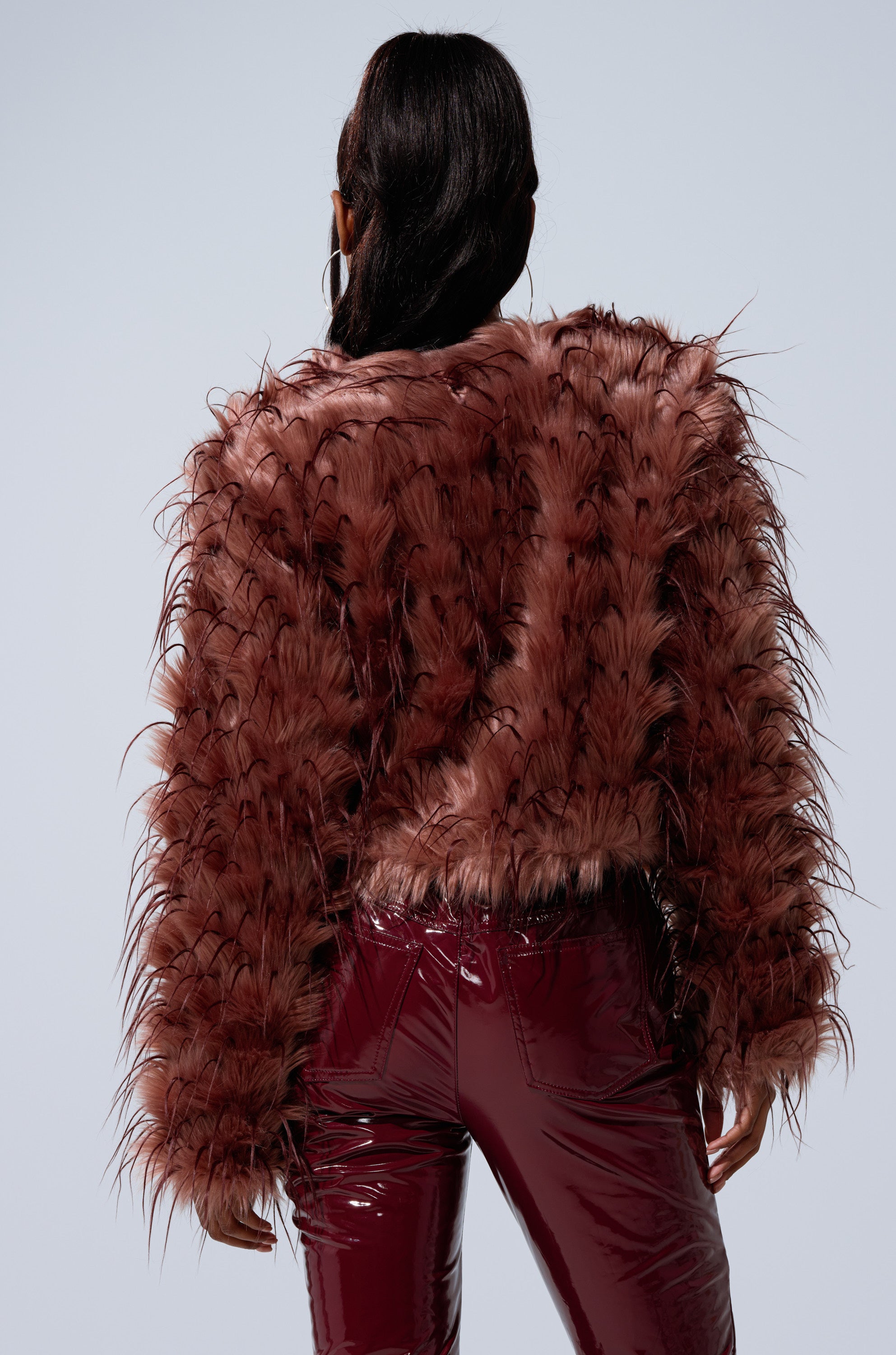 A person with long dark hair, seen from behind, wears the PIPER TEXTURED FAUX FUR COAT in maroon and matching shiny pants against a plain light background.