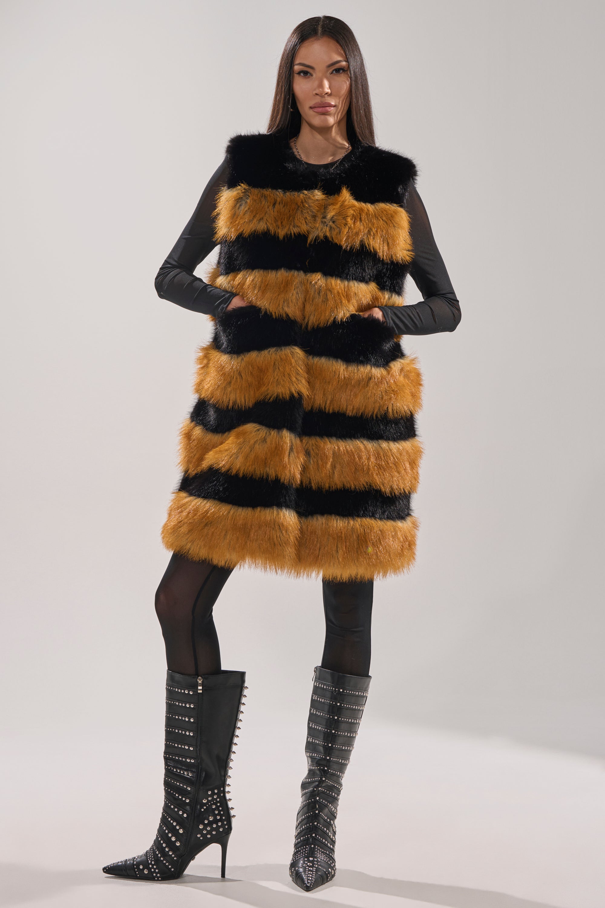 A woman stands confidently in the IN ANOTHER LIFE PANELED FUR VEST, a longline faux fur vest with orange and black stripes, paired with black tights and studded high-heeled boots against a light background.