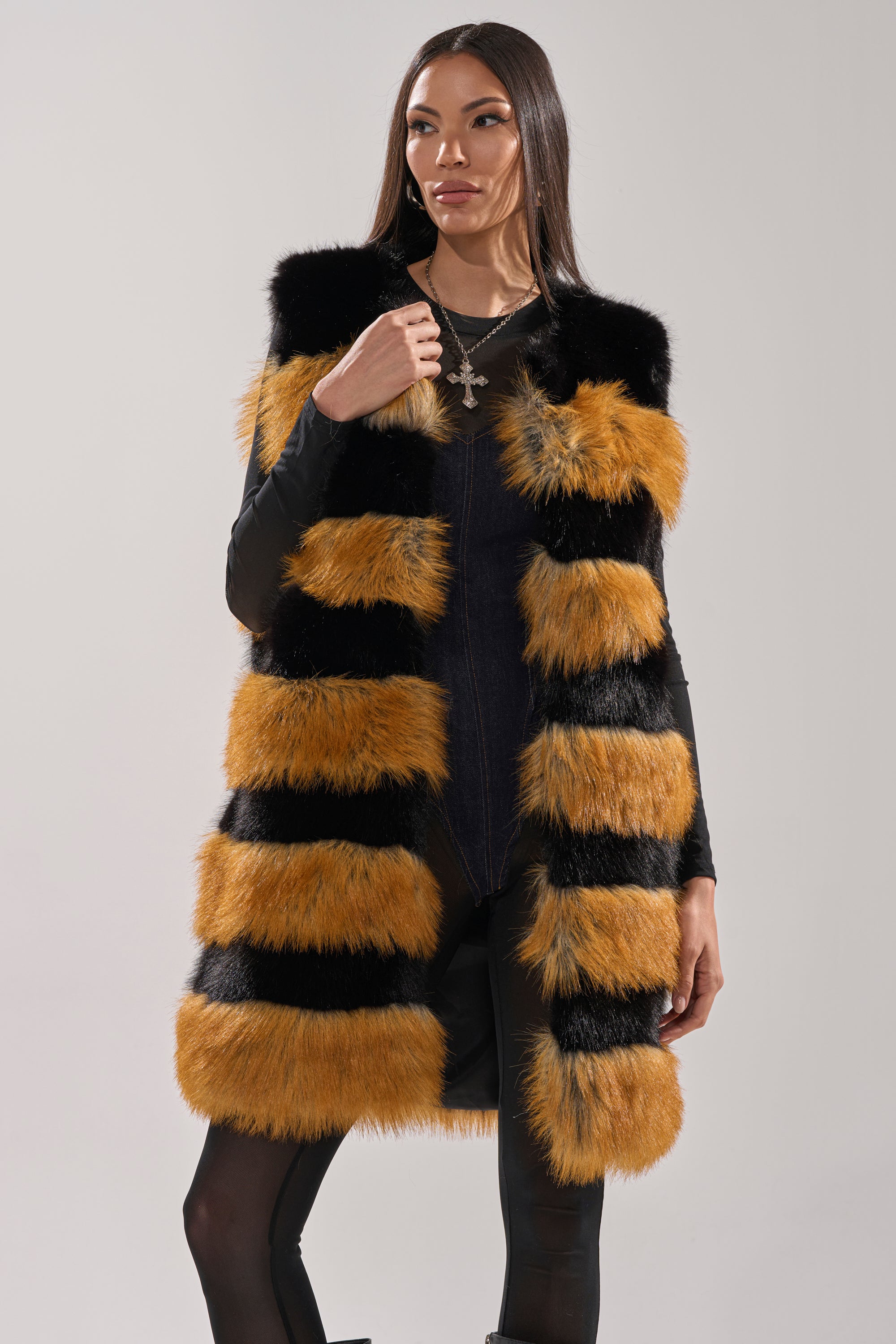 A woman models the IN ANOTHER LIFE PANELED FUR VEST, a longline faux fur piece with bold black and orange horizontal stripes, layered over a black outfit against a simple light background.