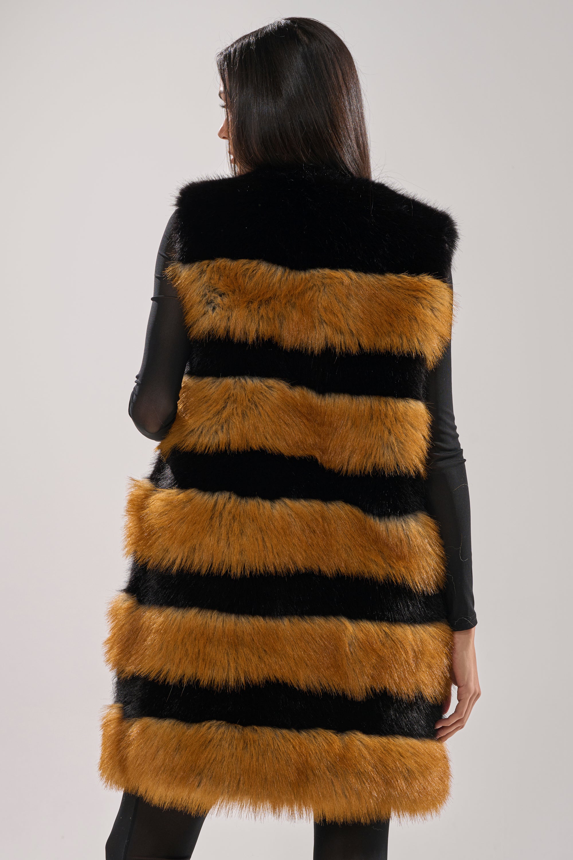 A person with long dark hair, seen from behind, wears the IN ANOTHER LIFE PANELED FUR VEST featuring sleeveless design and alternating horizontal black and orange-brown stripes, set against a plain light background.