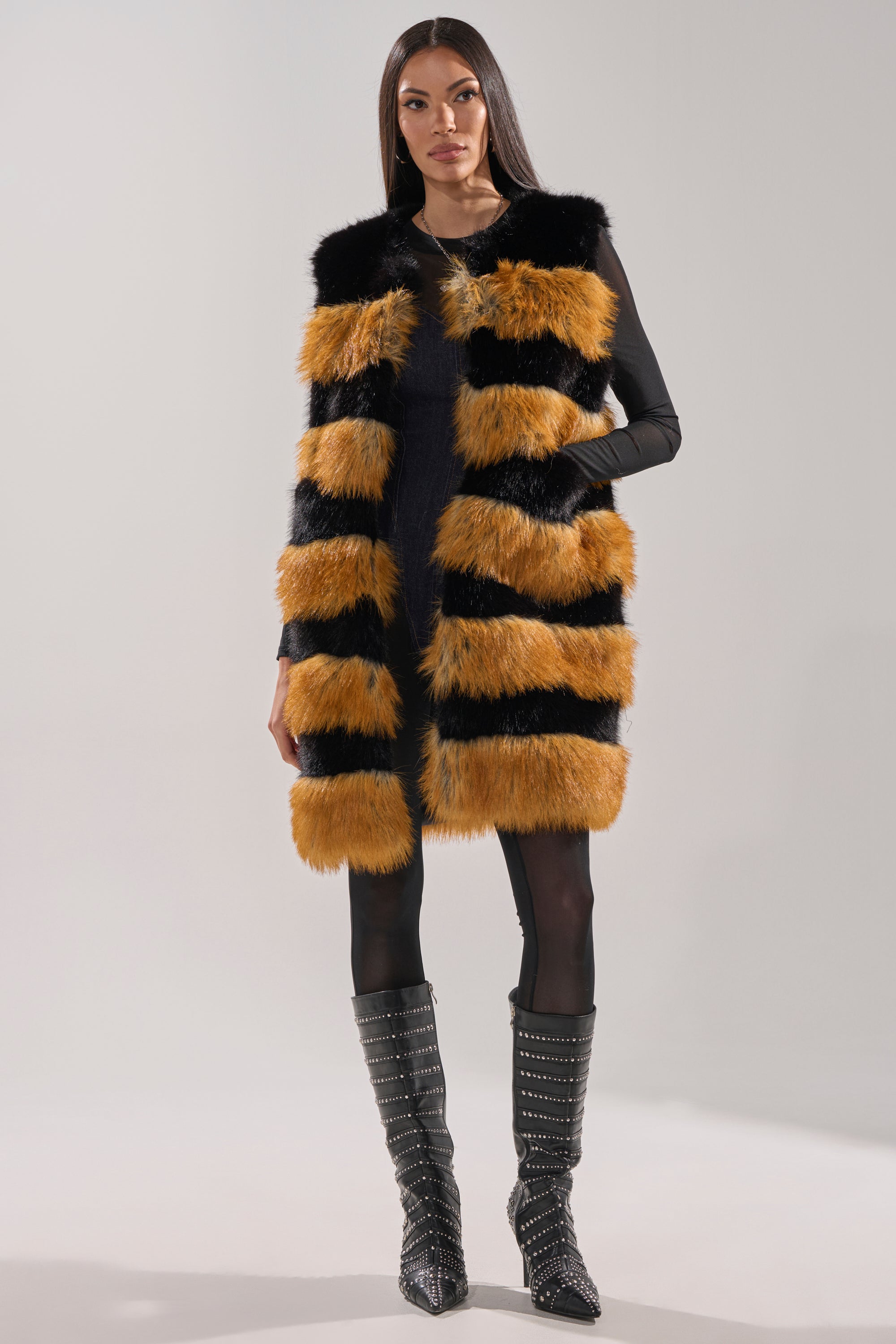 A woman models the IN ANOTHER LIFE PANELED FUR VEST, featuring a longline silhouette with black and orange panels, styled with a black dress, black tights, and tall black studded boots against a plain light gray background.