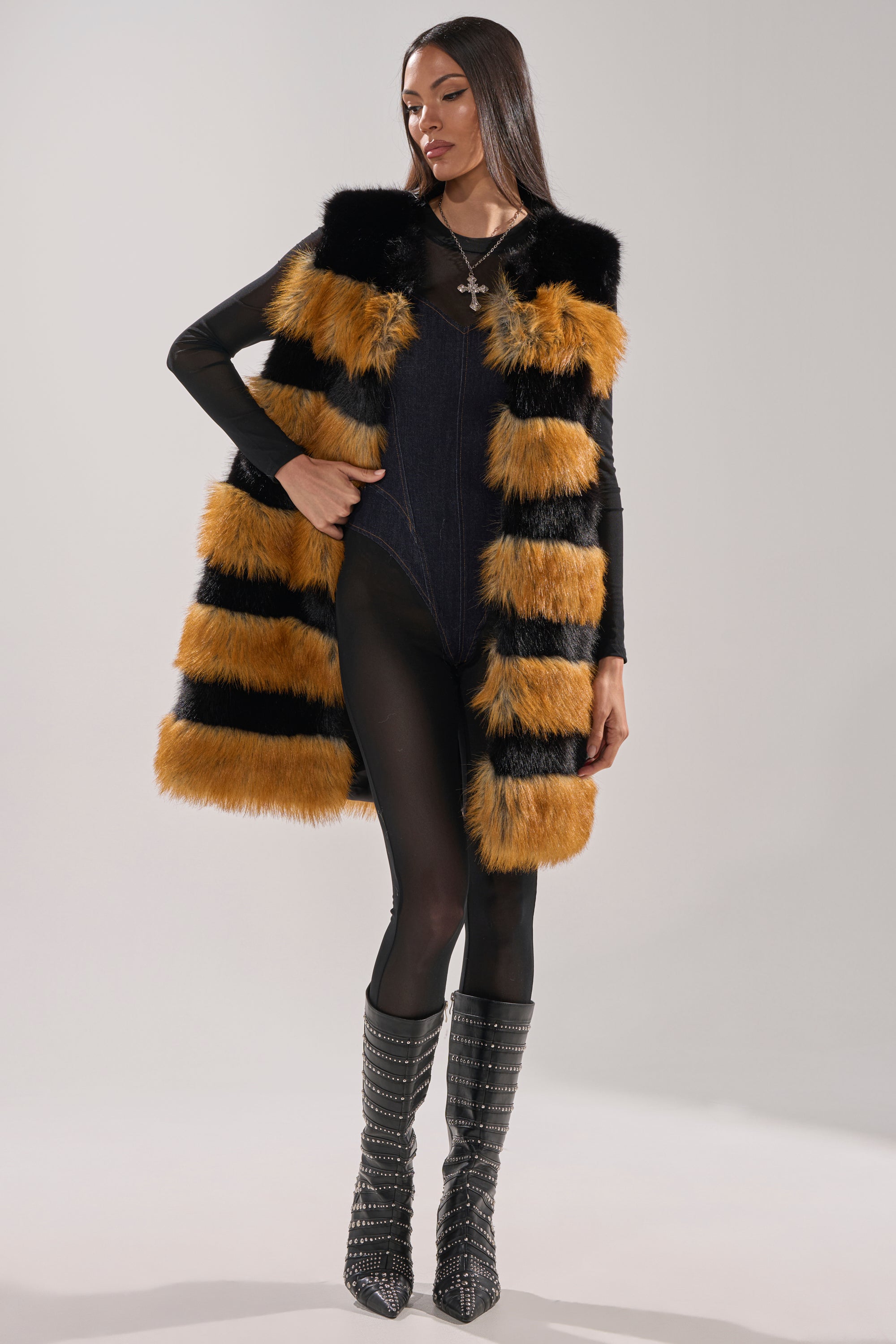 Wearing the IN ANOTHER LIFE PANELED FUR VEST with bold black and orange stripes, a woman poses in a black bodysuit and studded knee-high boots. Her long hair and necklace complement the striking, longline silhouette of her ensemble.