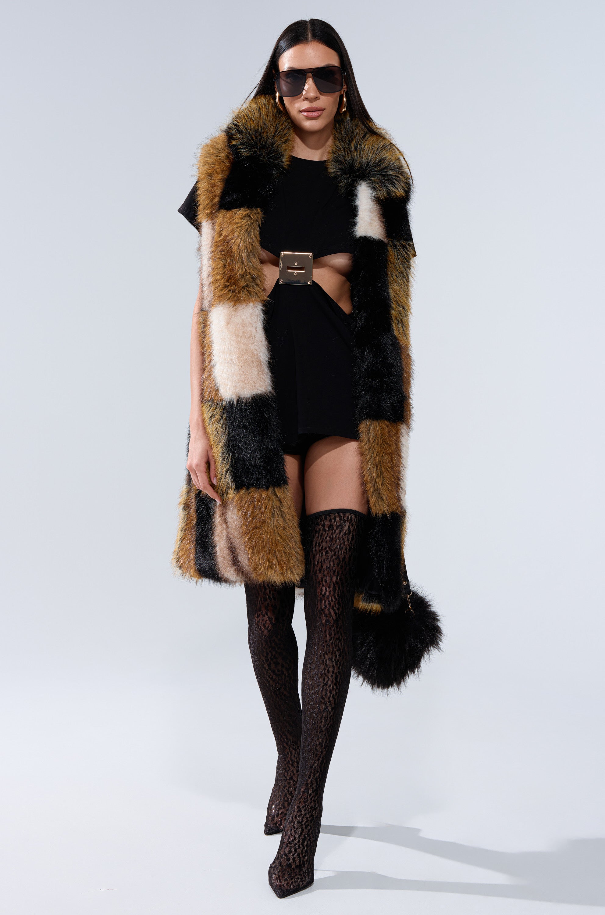 A model wearing oversized sunglasses, a black mini dress with cutouts, a wide belt, patterned thigh-high stockings, and holding a black fur bag showcases the CHECKMATE FAUX FUR VEST while walking forward.