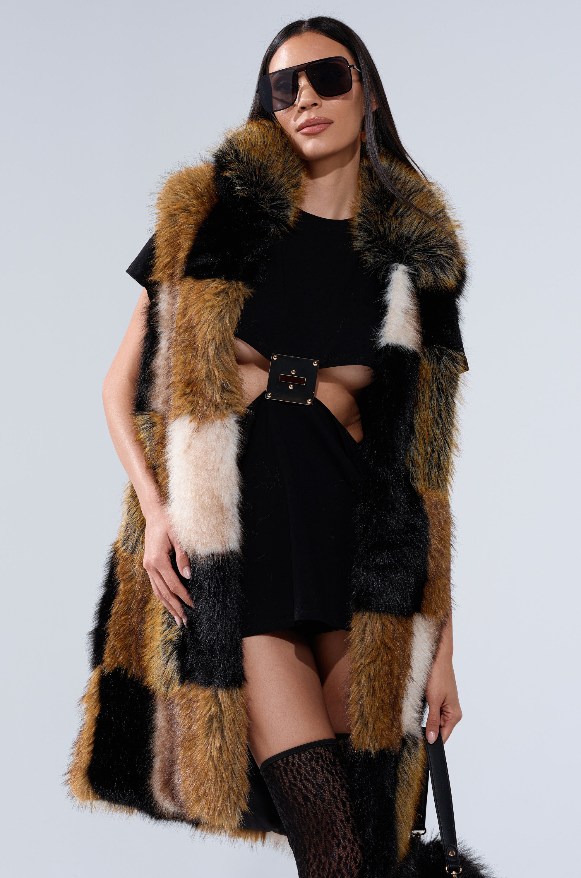 A woman models the CHECKMATE FAUX FUR VEST, styled over a black belted mini dress with sunglasses and thigh-high patterned boots, standing confidently against a plain background.