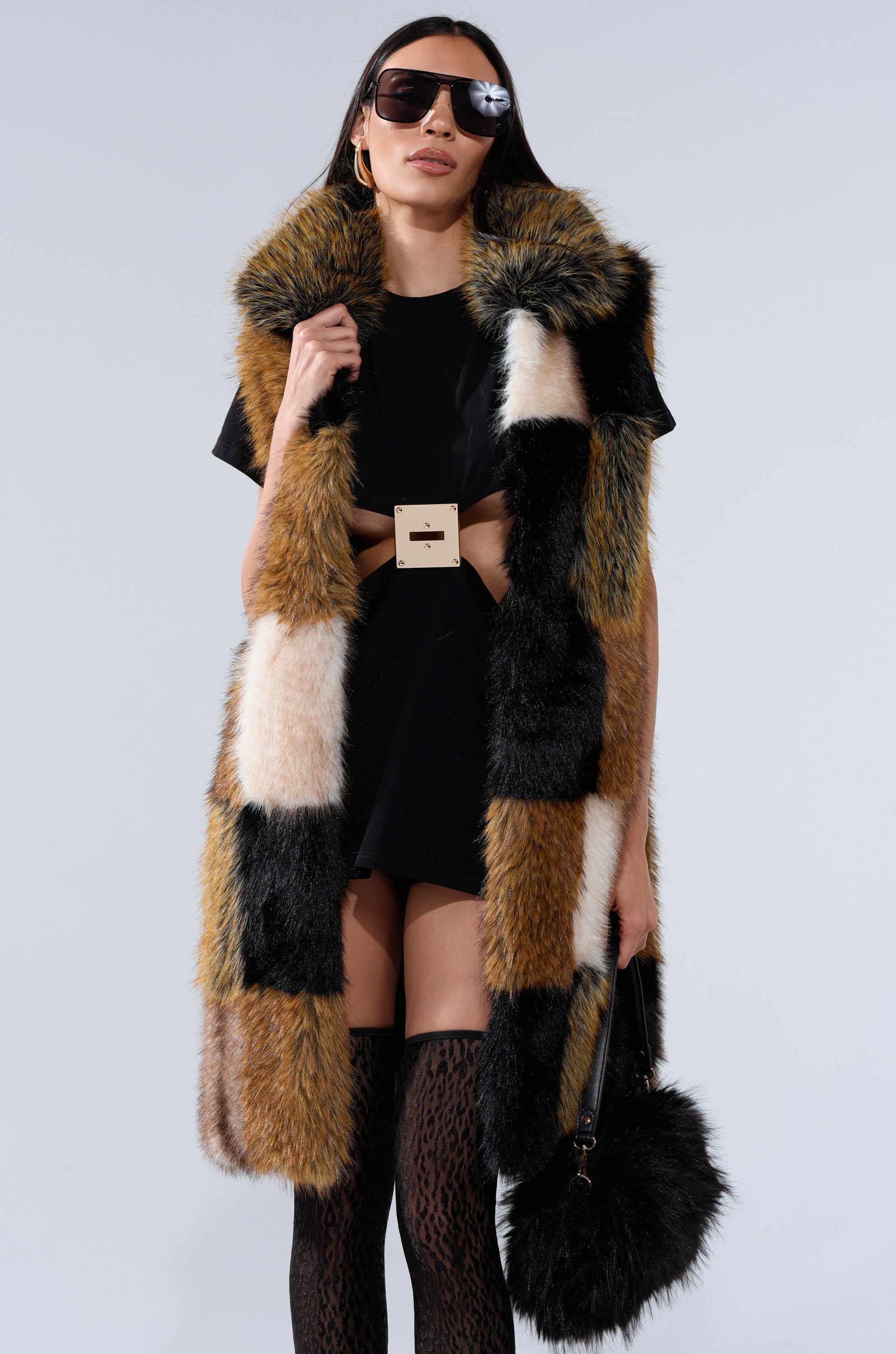 A woman poses confidently in the CHECKMATE FAUX FUR VEST over a black mini dress, with large sunglasses, a matching furry bag, and knee-high patterned black stockings.