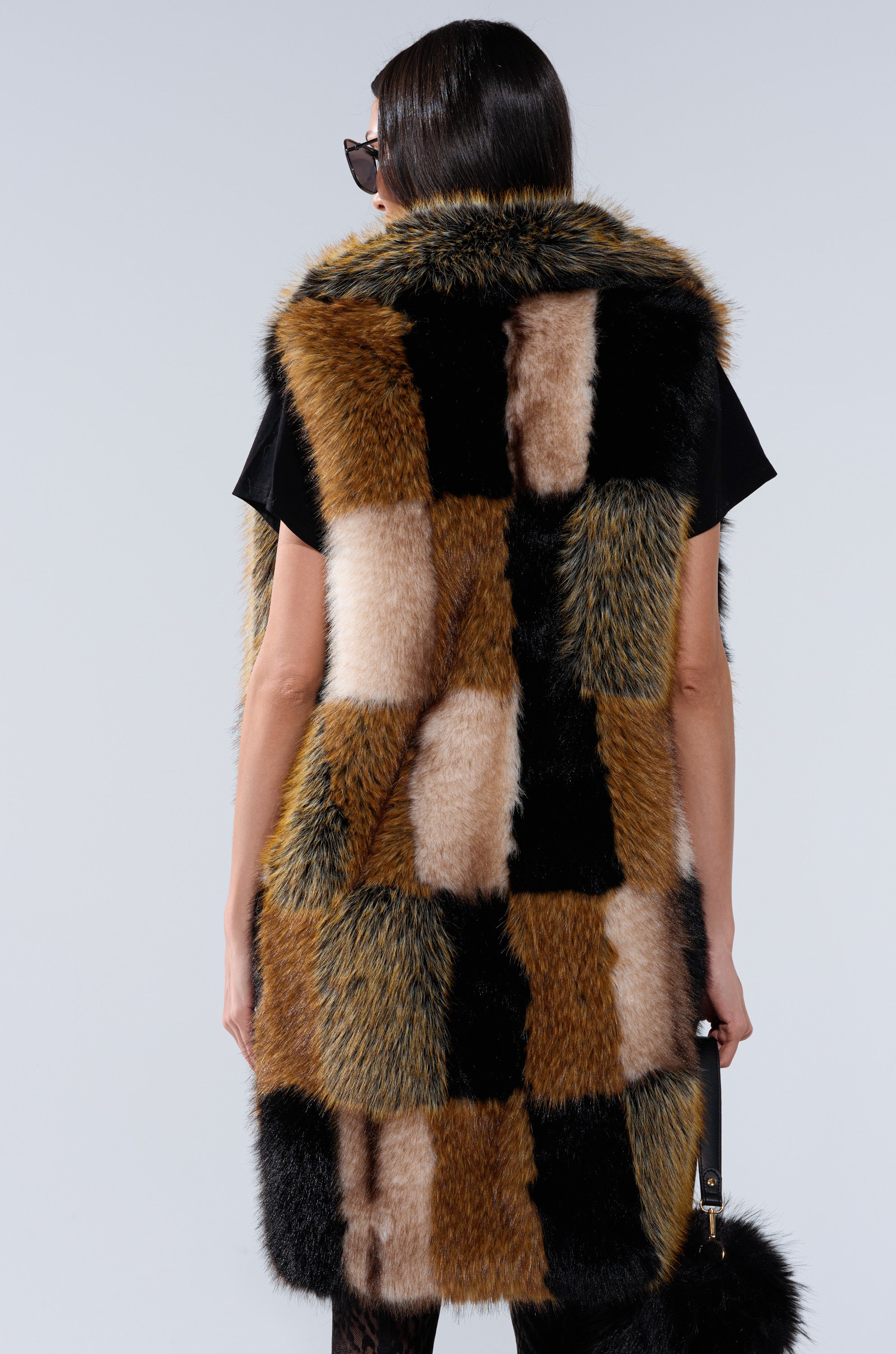 A person with straight dark hair and sunglasses is pictured from behind, wearing the CHECKMATE FAUX FUR VEST—a sleeveless piece with black, brown, tan, and cream checkered faux fur.