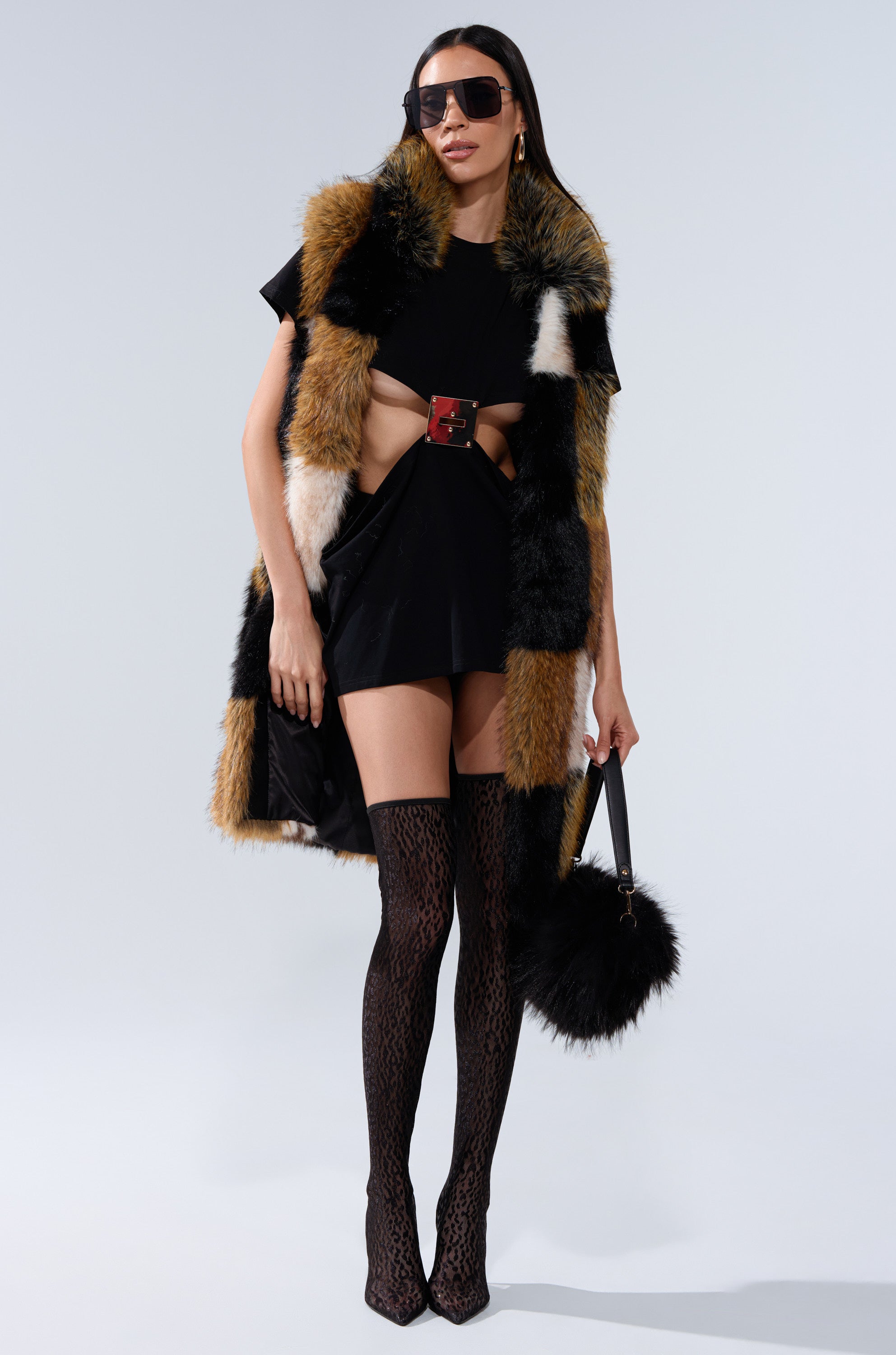 A woman in sunglasses, a black mini dress, wide belt, patterned thigh-high boots, holding a black furry handbag showcases her bold look featuring the CHECKMATE FAUX FUR VEST against a plain background.