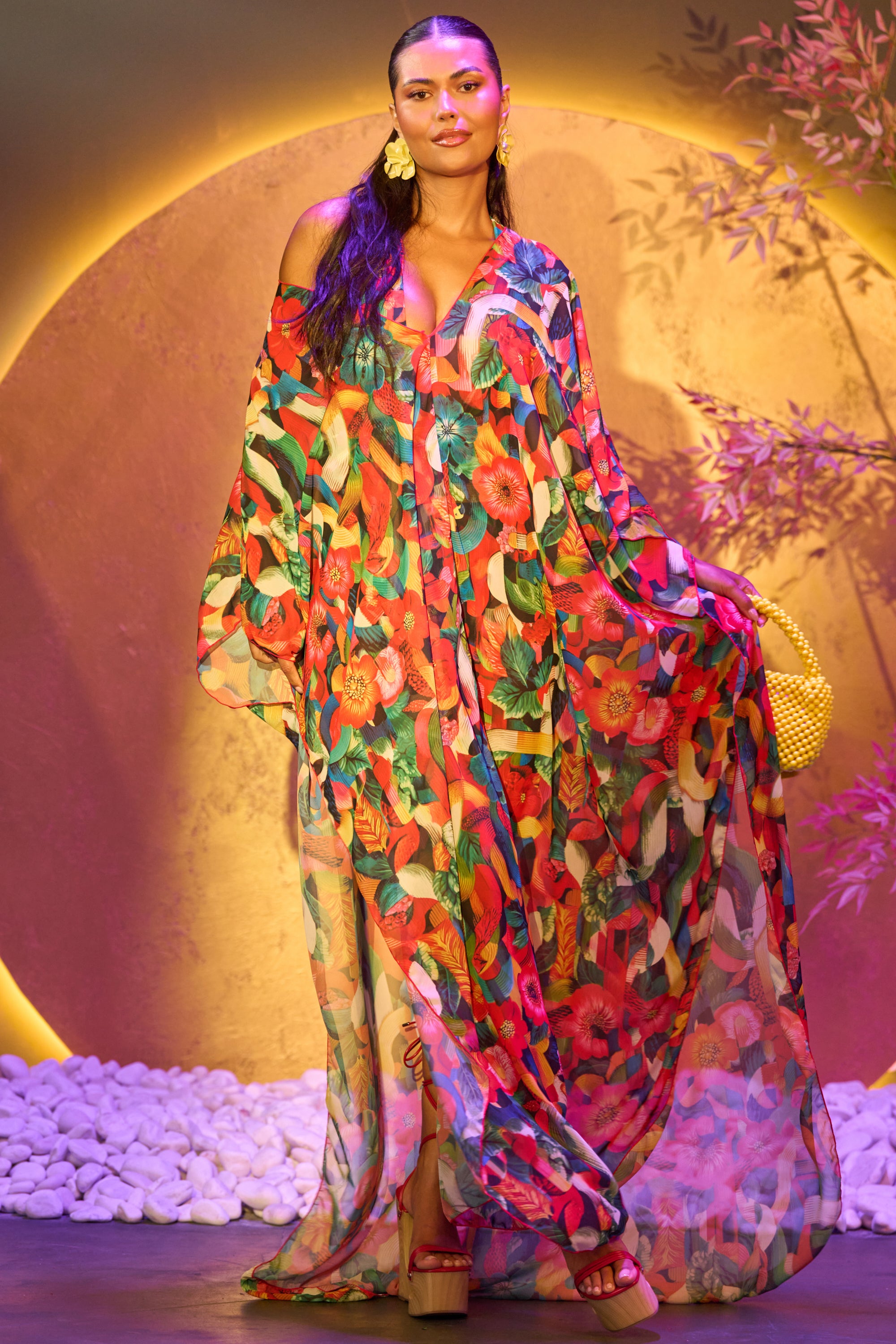 A woman models the SWEET LIFE KAFTAN, a flowing, colorful floral dress with long, wide sleeves. She accessorizes with statement earrings, platform sandals, and a woven bag against a large yellow circle and artistic foliage backdrop. 1
