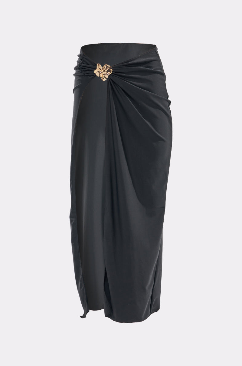 The TO THE YACHT SWIM COVER SKIRT is a long black skirt with a draped design, featuring a gathered knot at the waist with a gold brooch. Its soft-sheen fabric flows smoothly to ankle length. 3