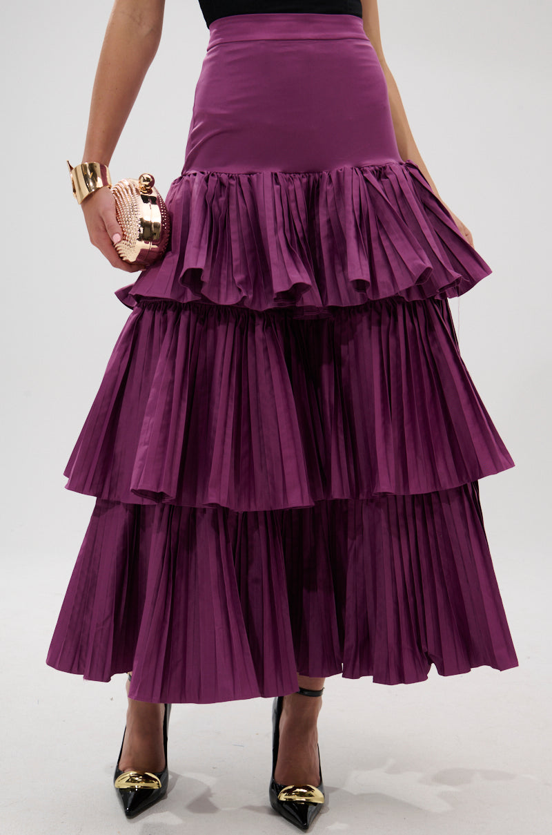 A person showcases the DANCE AROUND WITH ME PLEATED MAXI SKIRT IN PLUM, holding a round metallic clutch and wearing black pointed heels with gold tips. The image is cropped to highlight only the lower torso and legs.