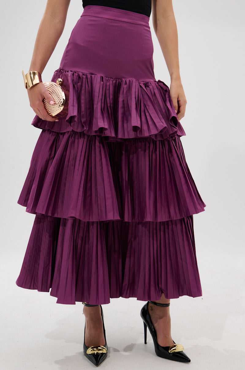 A person wears the DANCE AROUND WITH ME PLEATED MAXI SKIRT IN PLUM, paired with black pointed heels featuring gold tips, and holds a round metallic pink clutch. Only the lower half and part of the upper body are visible.