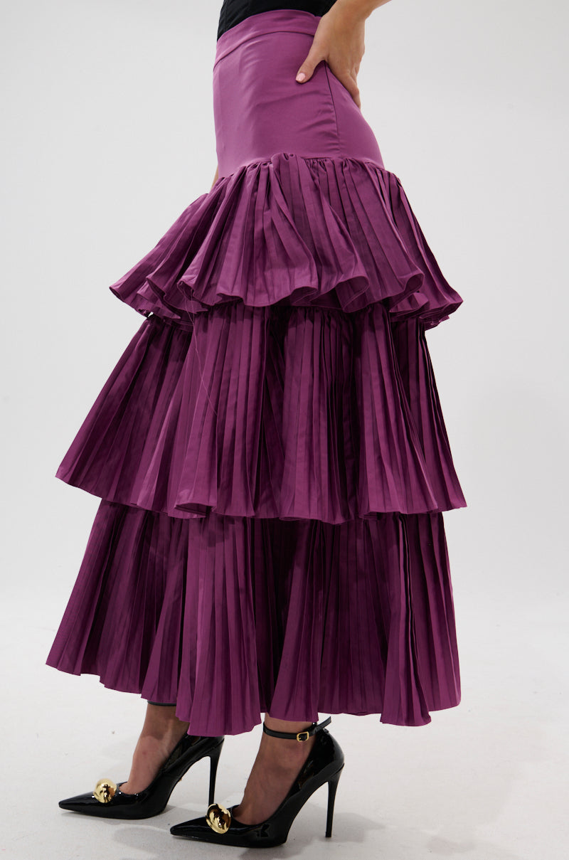 Wearing the DANCE AROUND WITH ME PLEATED MAXI SKIRT IN PLUM and black pointed heels with gold embellishments, a person stands against a plain white background.