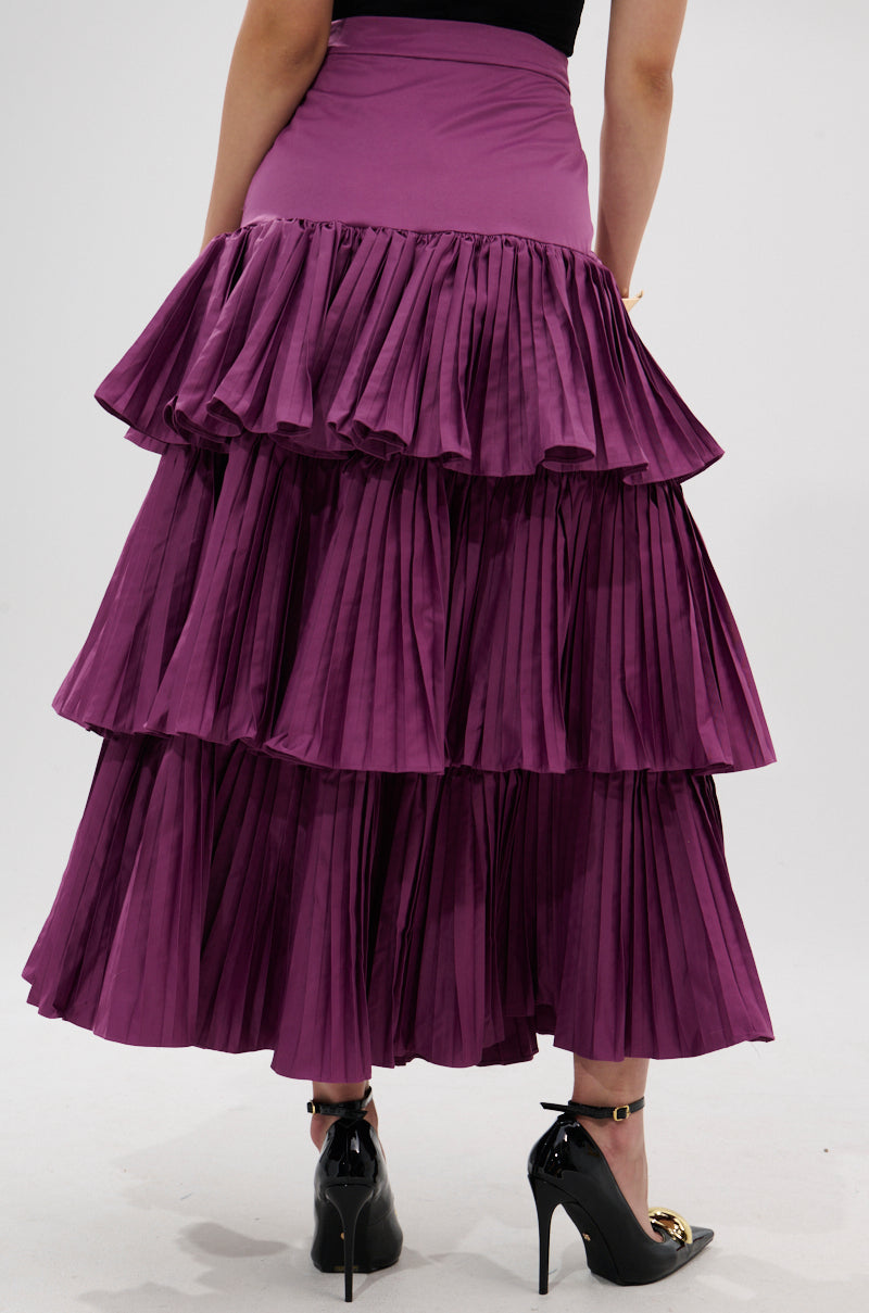 A person wearing the DANCE AROUND WITH ME PLEATED MAXI SKIRT IN PLUM and black high-heeled shoes is photographed from the back against a plain light background.