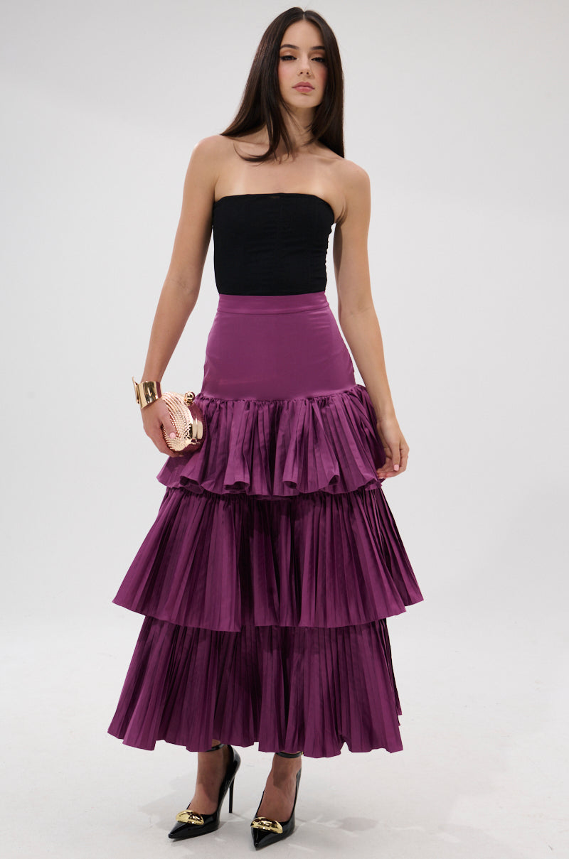 A woman stands against a plain background wearing a black strapless top and the DANCE AROUND WITH ME PLEATED MAXI SKIRT IN PLUM, styled with a gold clutch, black heels with gold accents, and a matching gold bracelet.