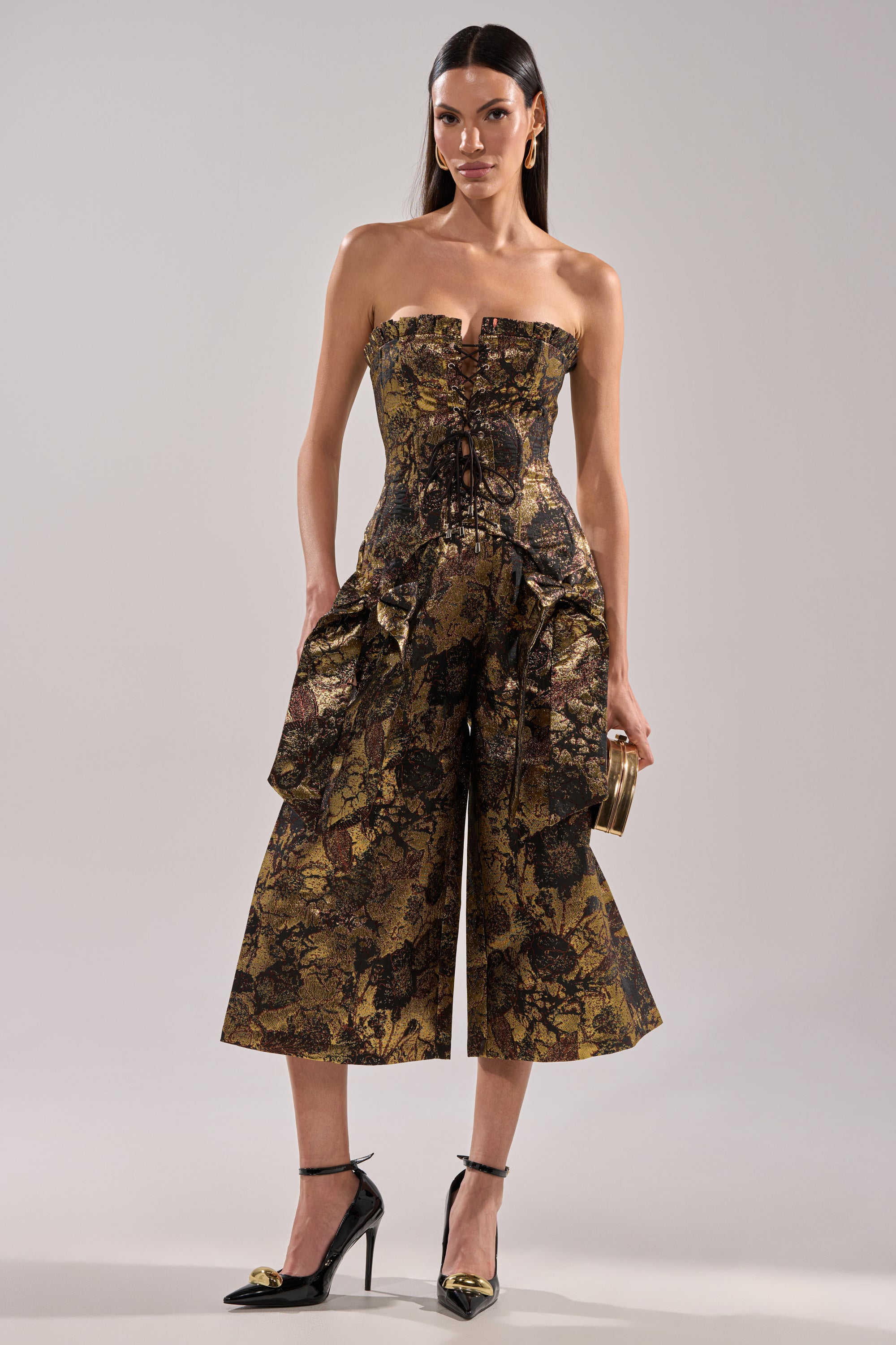 Wearing the LOTUS BROCADE CAPRI jumpsuit with a strapless, fitted bodice and wide-leg cropped pants, a woman pairs her look with black pointed-toe ankle strap heels and carries a small clutch.