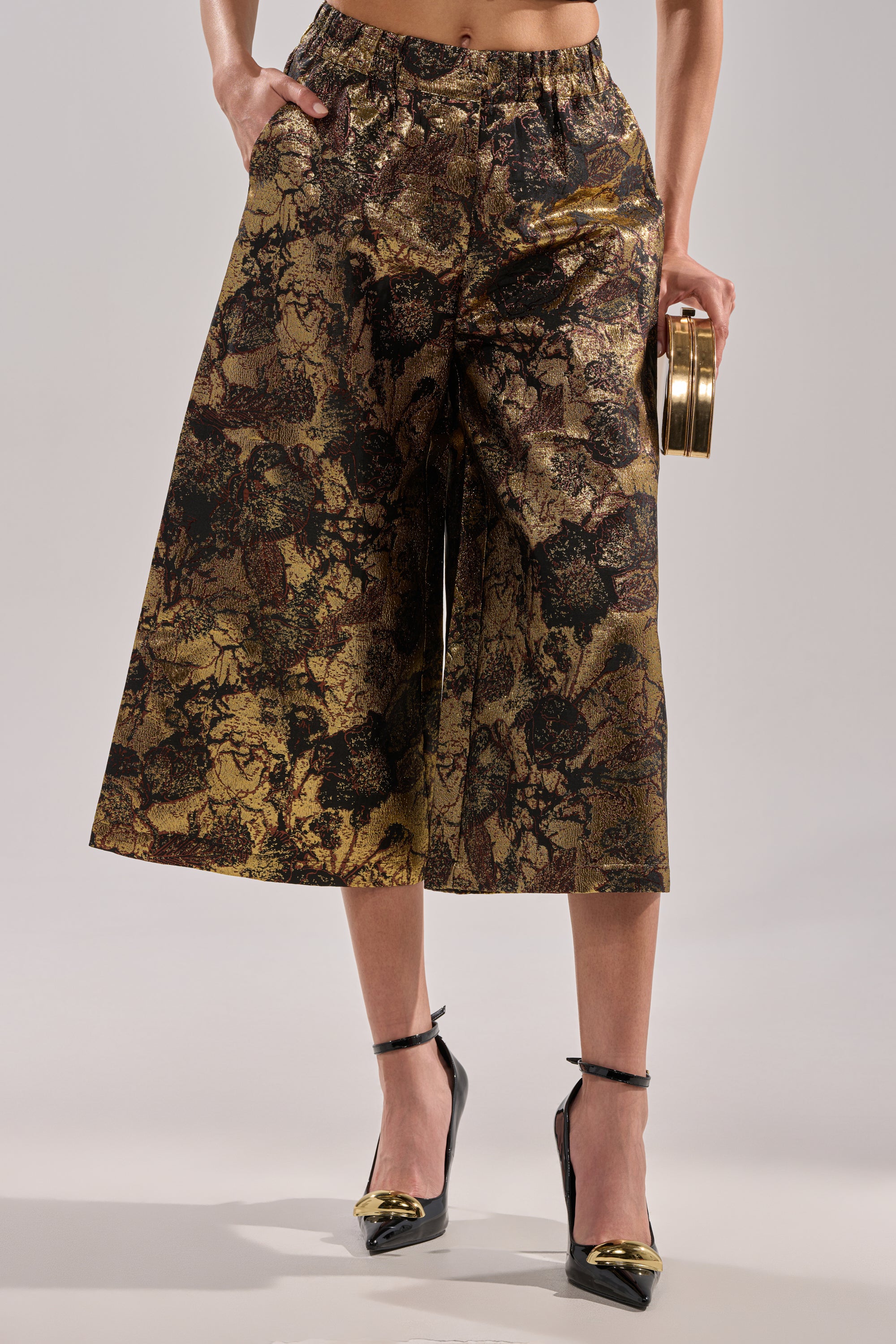 A woman models the LOTUS BROCADE CAPRI in metallic gold and black floral, styled with black ankle-strap heels with gold tips and a gold round clutch. The neutral background focuses on her lower body.